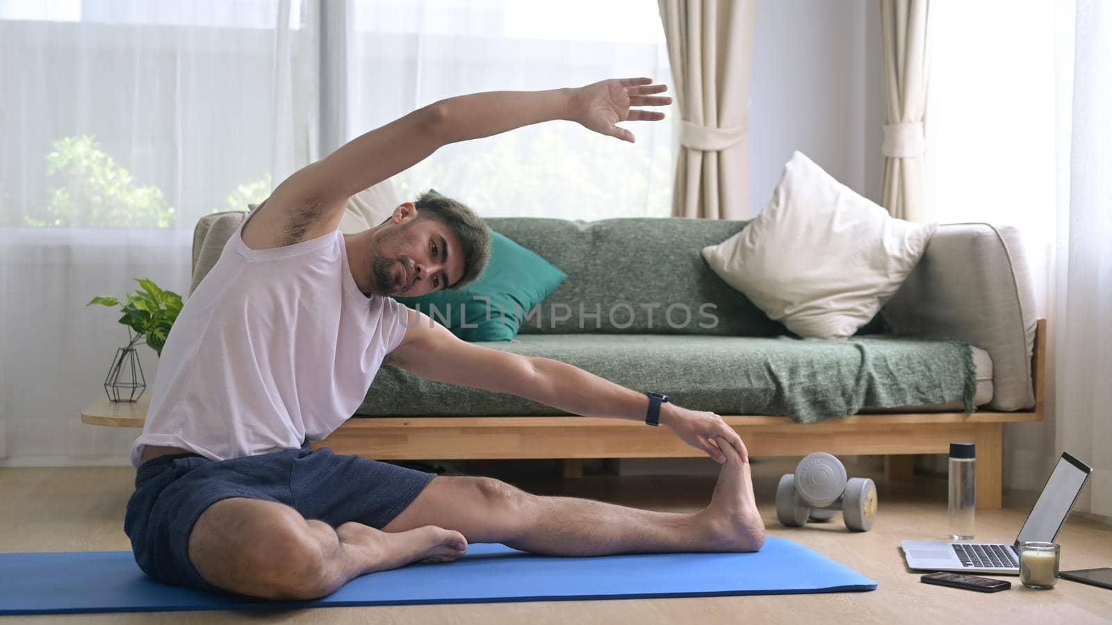 Young man in sports clothes is warming up, stretching while sitting on mat at home by prathanchorruangsak
