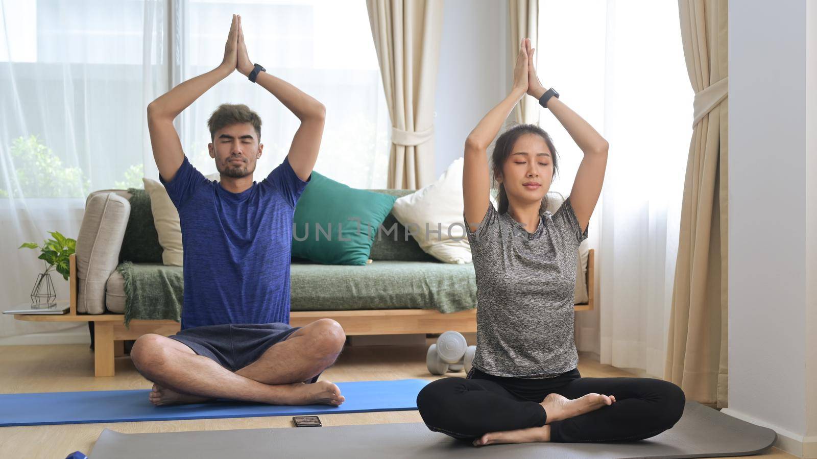 Horizontal view of healthy young couple sitting in lotus position while practicing yoga in living room.