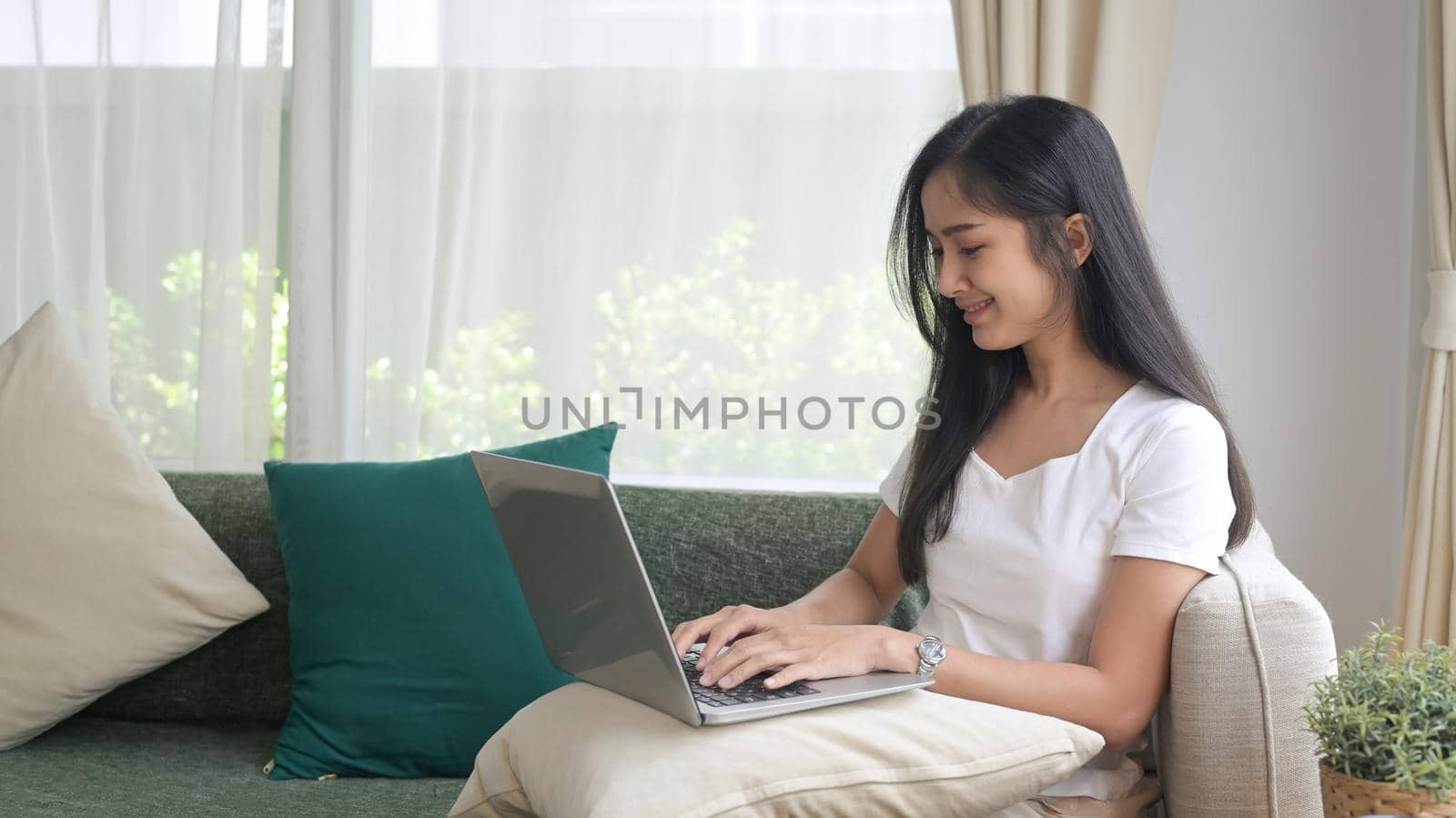 Smiling young woman in casual clothes resting on sofa and and using laptop, surfing internet, checking email.