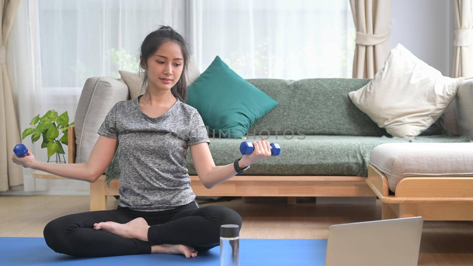 Active young woman in sportswear exercising with dumbbells and watching fitness lessons online on laptop by prathanchorruangsak