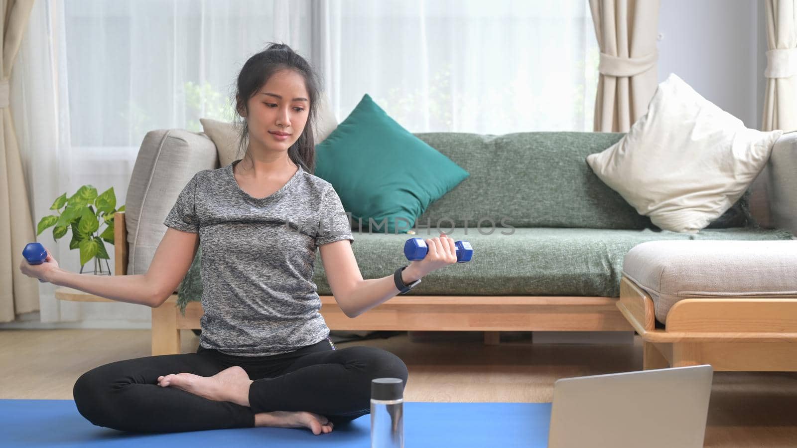 Active young woman in sportswear exercising with dumbbells and watching fitness lessons online on laptop.