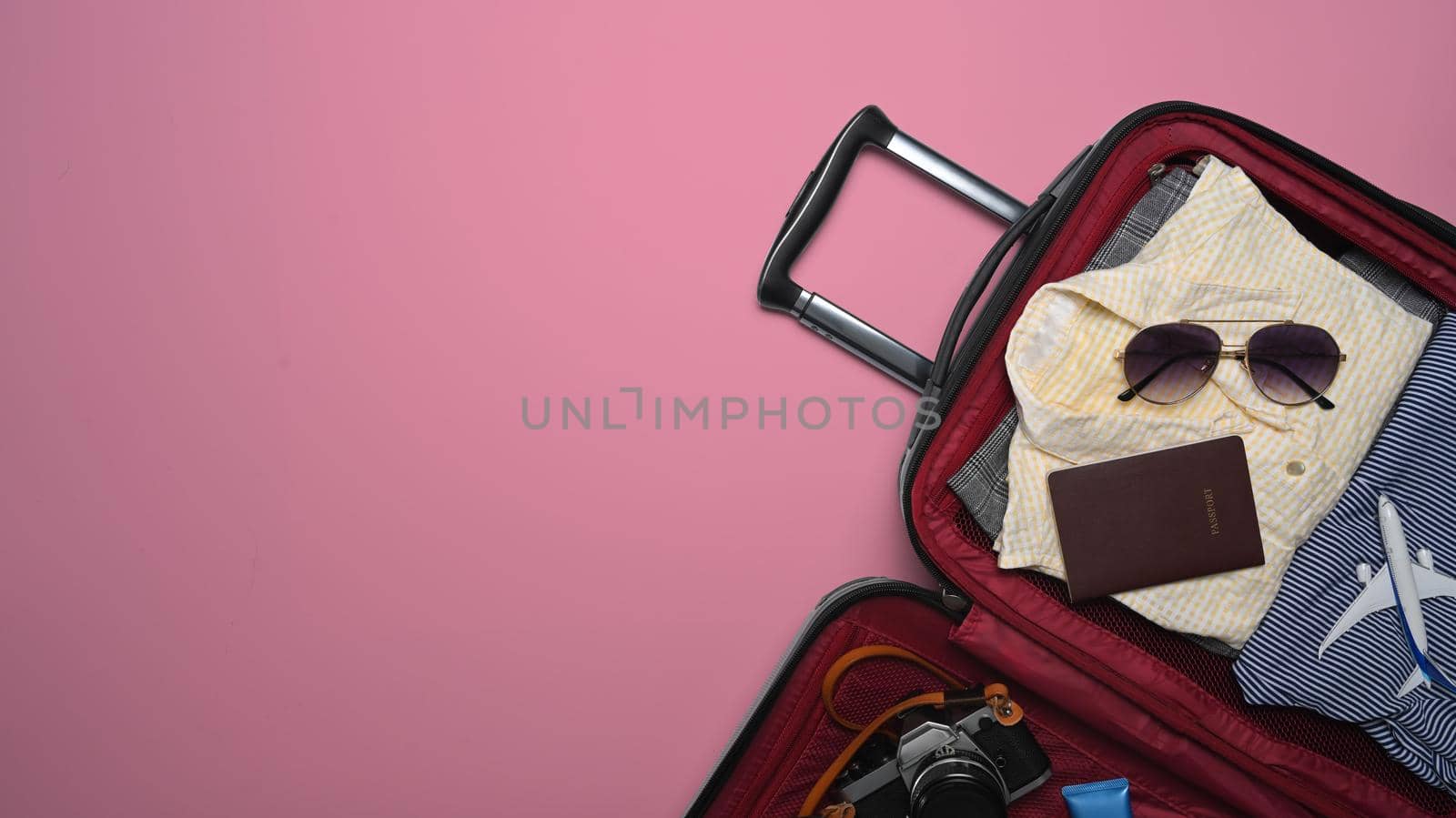 Open suitcase with sunglasses, camera, passport and clothes on pink background. Preparing for the summer vacations.