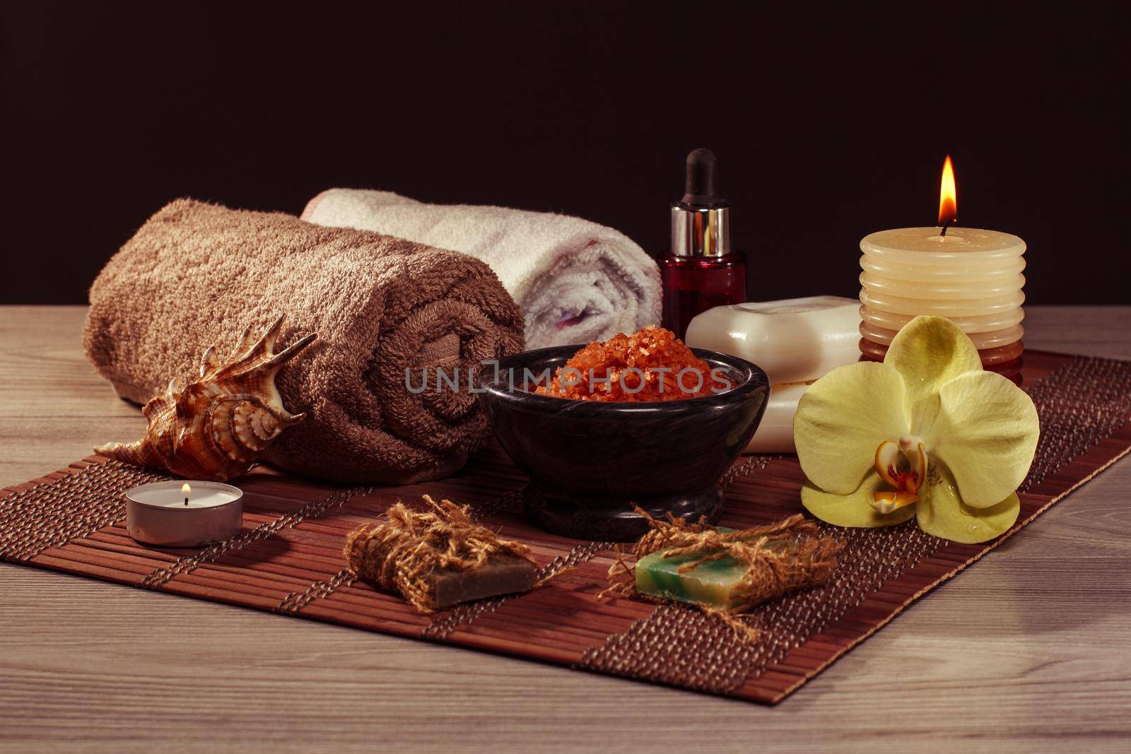 Spa and wellness setting with orchid flower, bowl with sea salt, seashell, bottle with aromatic oil, soap, candles and towel on bamboo napkin