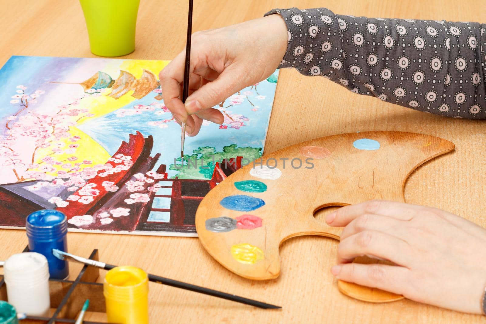 Woman's hand painting Japanese landscape indoors on the wooden desk with palette, paints and paintbrushes