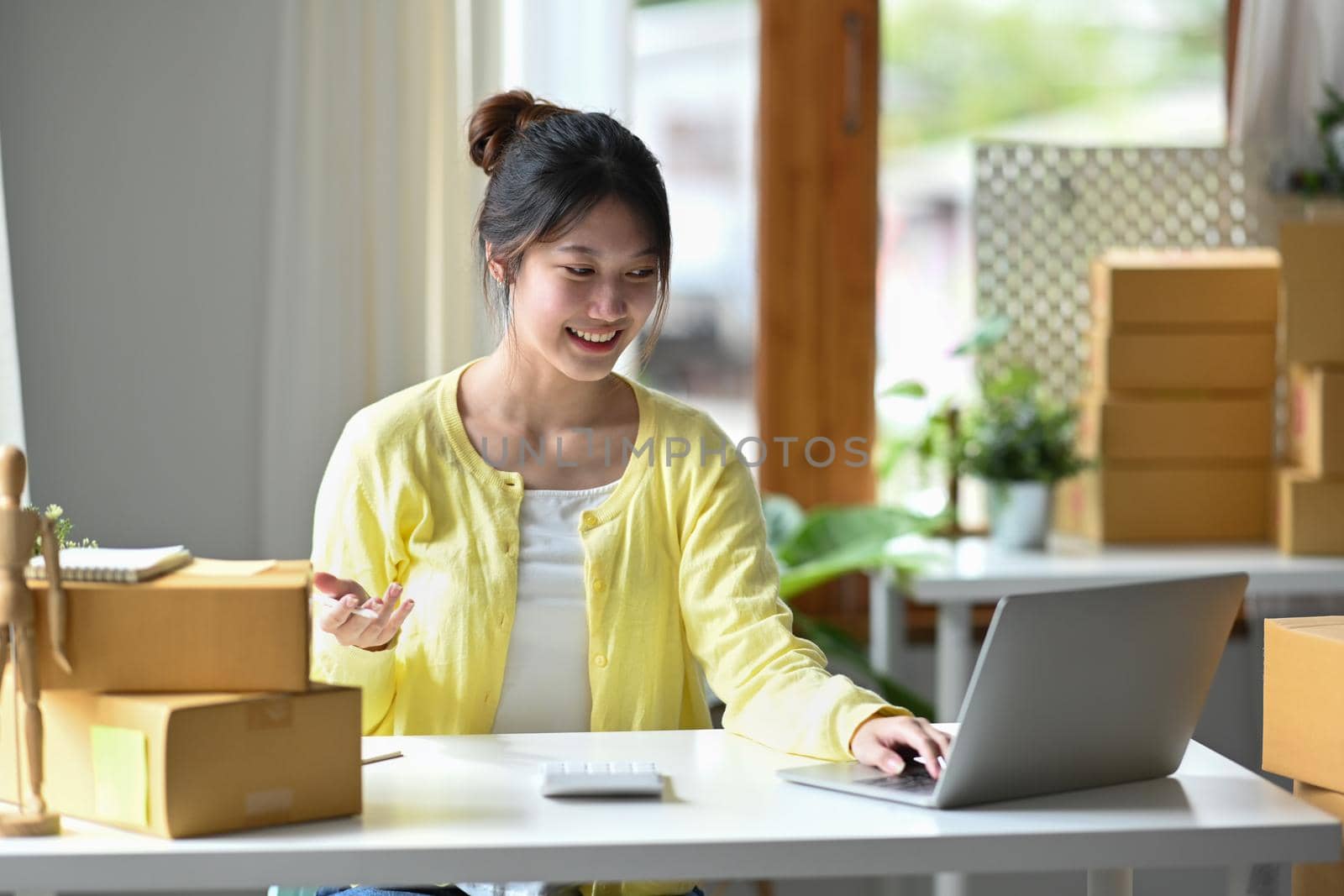 Smiling young female entrepreneur checking online order on digital tablet and preparing parcel boxes for delivery to customers.