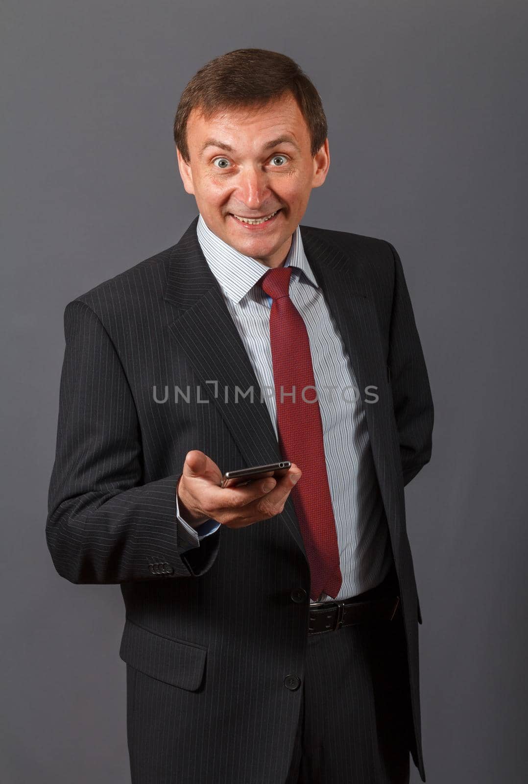 Elegant handsome mature businessman standing in front of a gray background in a studio wearing a nice suit holding a sell phone and having funny facial expression.