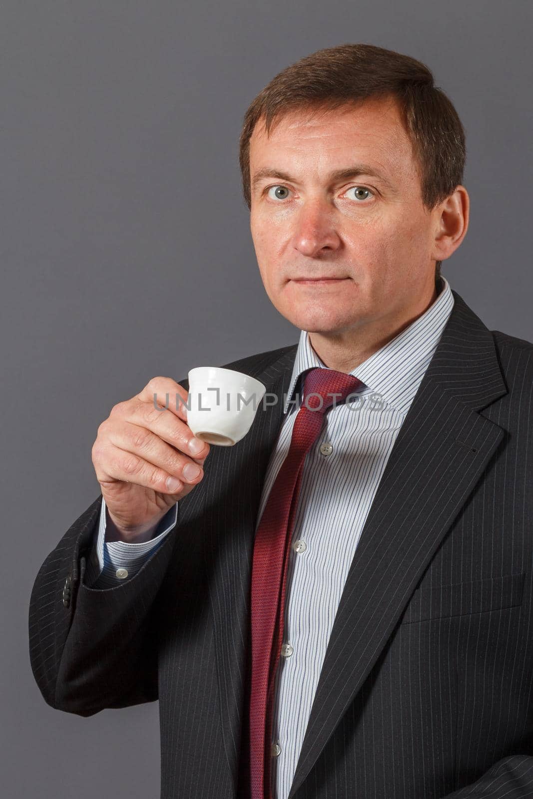 Confident and friendly elegant handsome mature businessman standing in front of a gray background in a studio taking a cup of coffee
