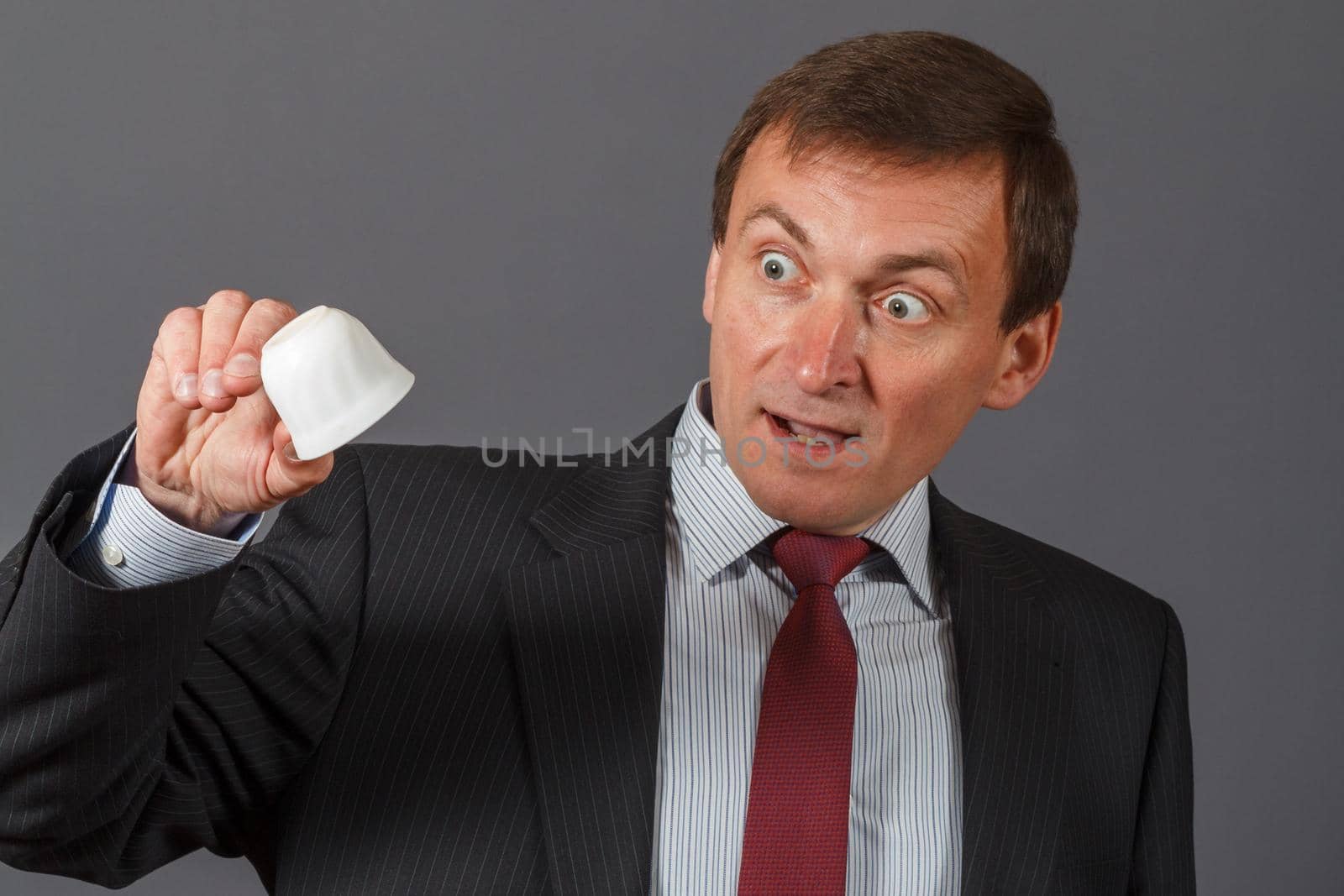 Elegant handsome mature businessman standing in front of a gray background taking an empty cup of coffee and having surprised expression.