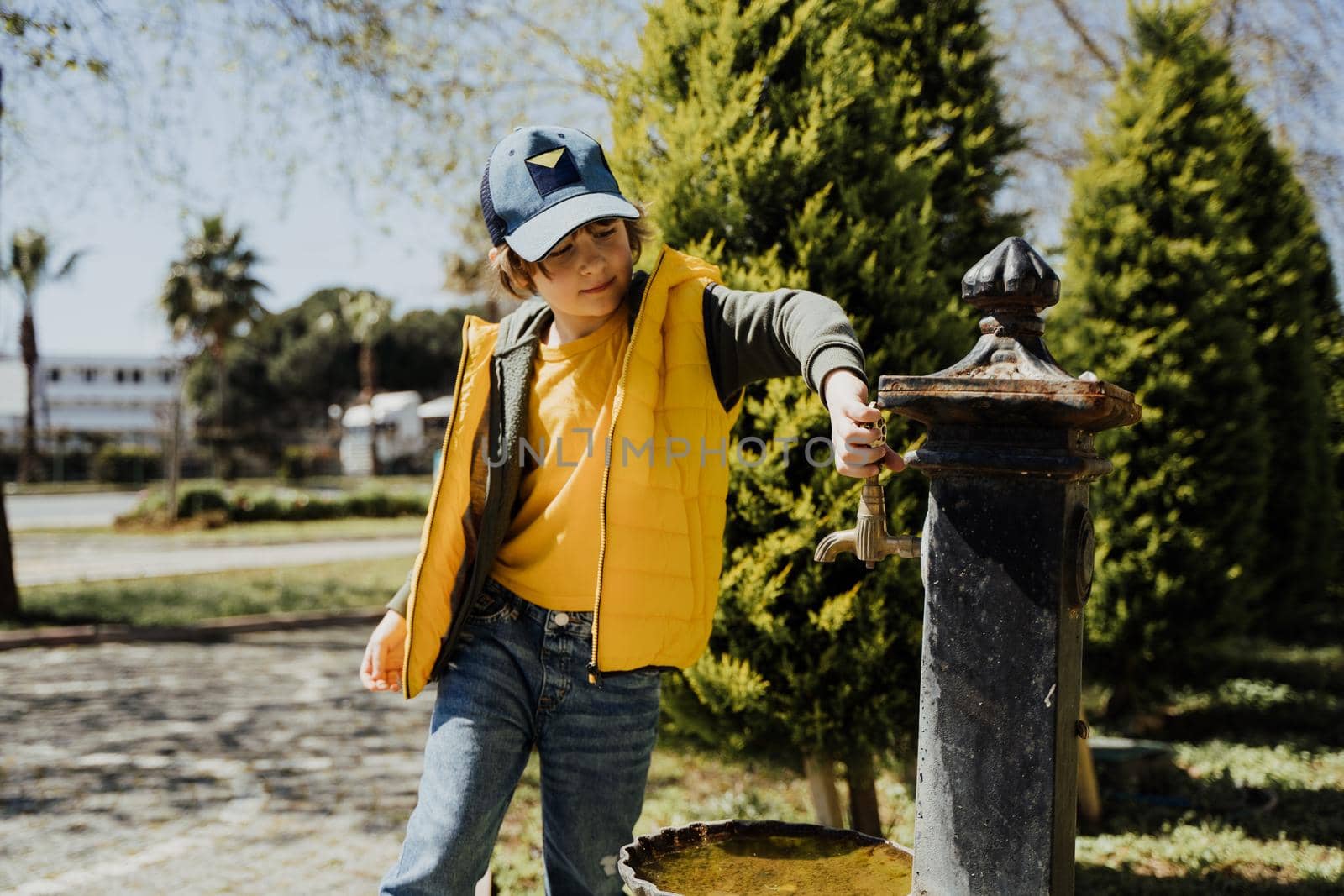 Kid schoolboy in casual clothing washing his hands in the street old fashioned drinking water fountain. Boy in blue jeans and yellow vest playing with water from drinking sprinkler in city park.