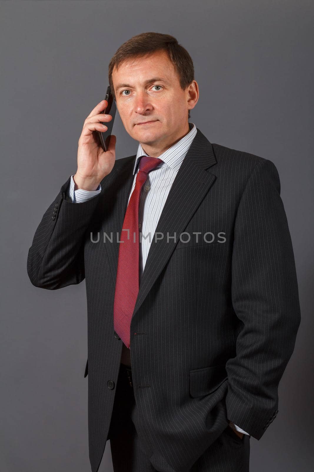 Confident elegant handsome mature businessman standing in front of a gray background in a studio wearing a nice suit is speaking on the sell phone