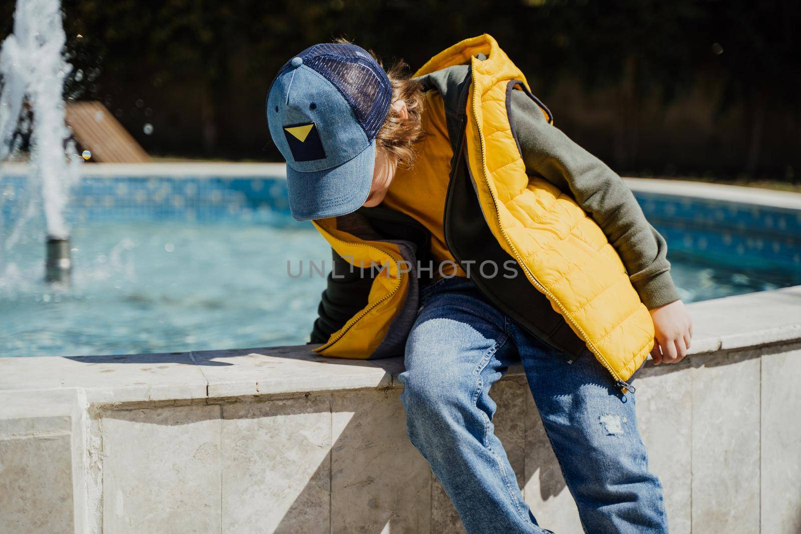 Schoolboy sitting on a fountain in public park during spring sunny day playing with water. Kid in yellow vest chilling in park with water sprinkling in the background. Summer holidays outside.