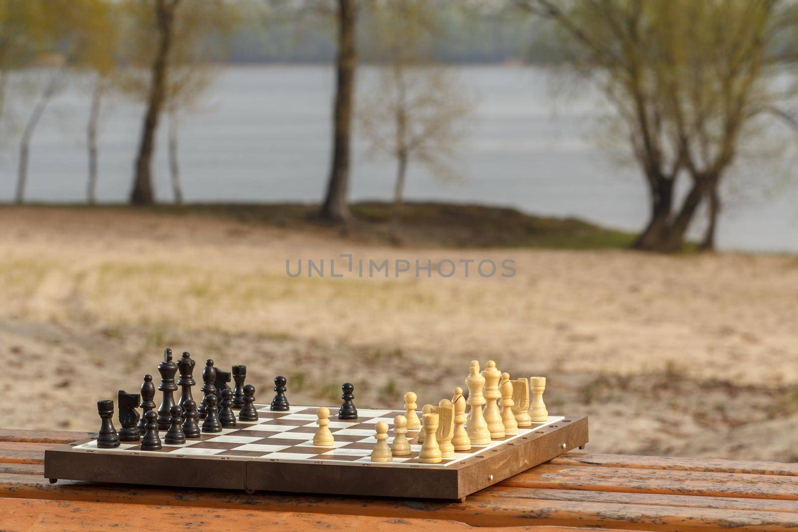 One chess pieces staying against black chess pieces Chess board with chess pieces on wooden bench with river embankment background