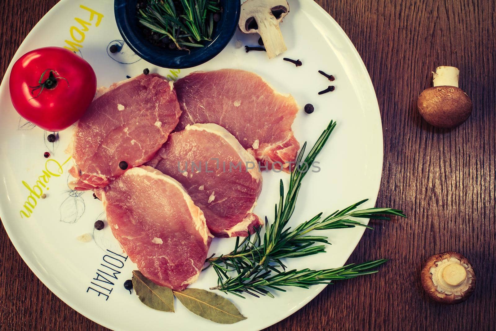 Pieces of raw pork steak with spices and herbs rosemary, mushrooms, tomato, salt and pepper on a white plate, top view