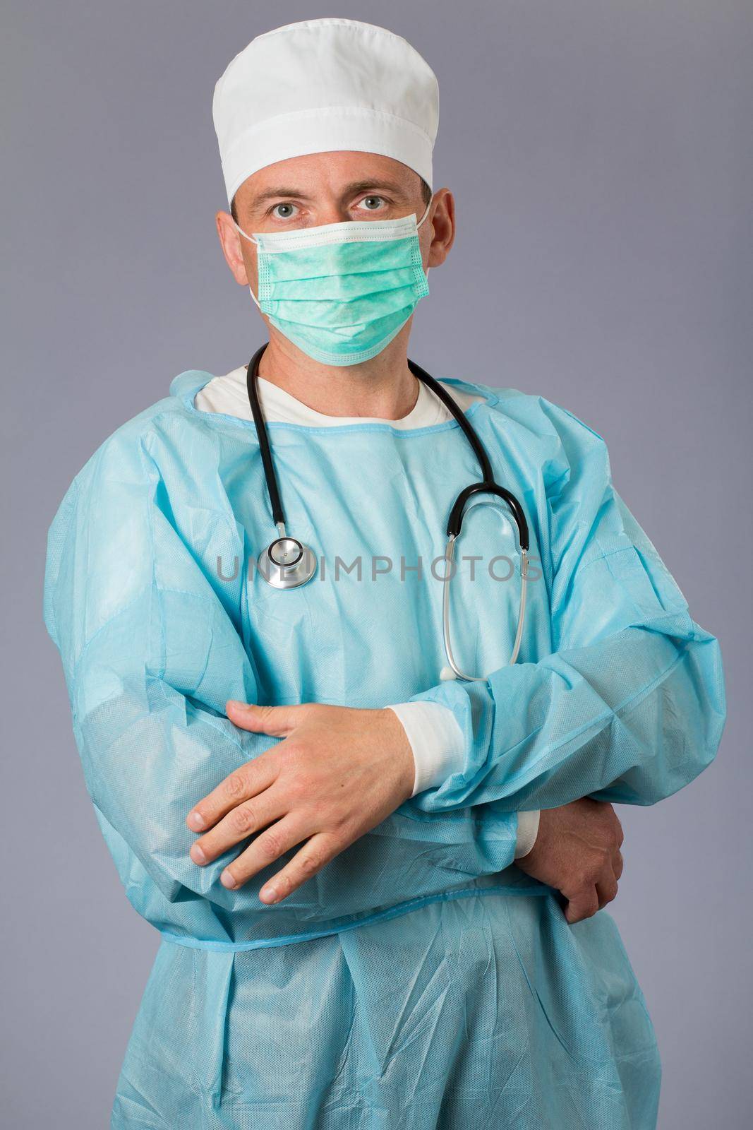 Medical doctor dressed in a medical gown with stethoscope and face mask standing with arms folded. Gray background