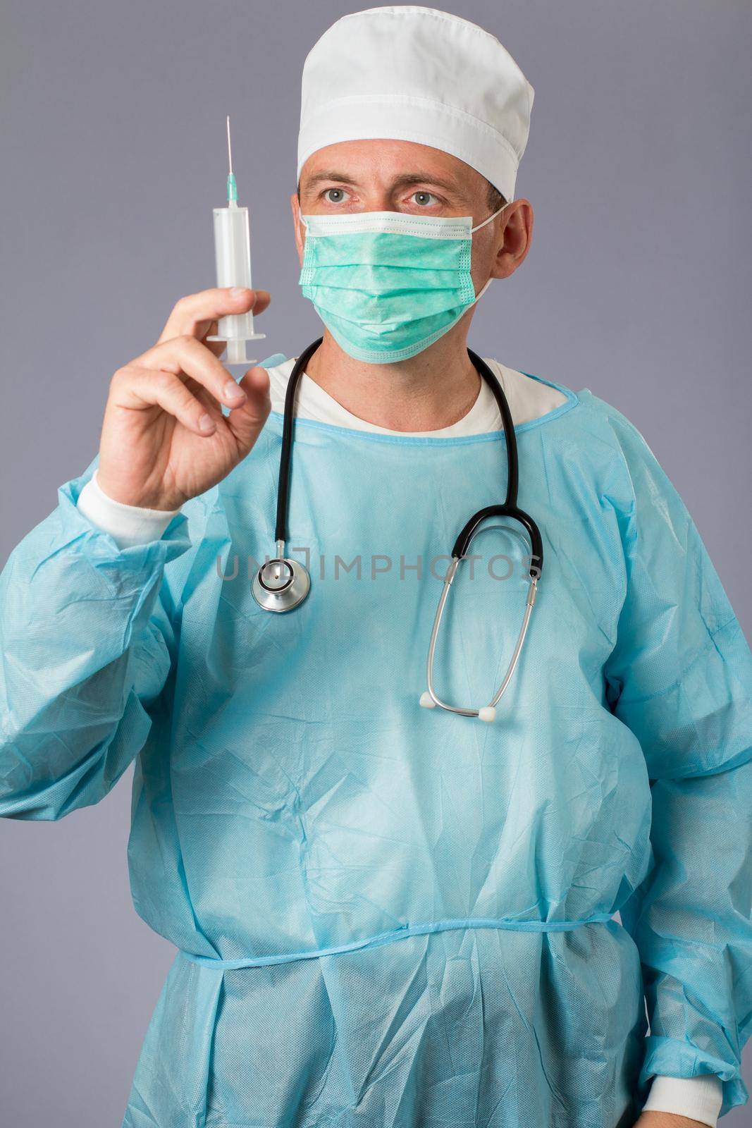 Medical doctor dressed in a medical gown with stethoscope and face mask holding a syringe. Gray background