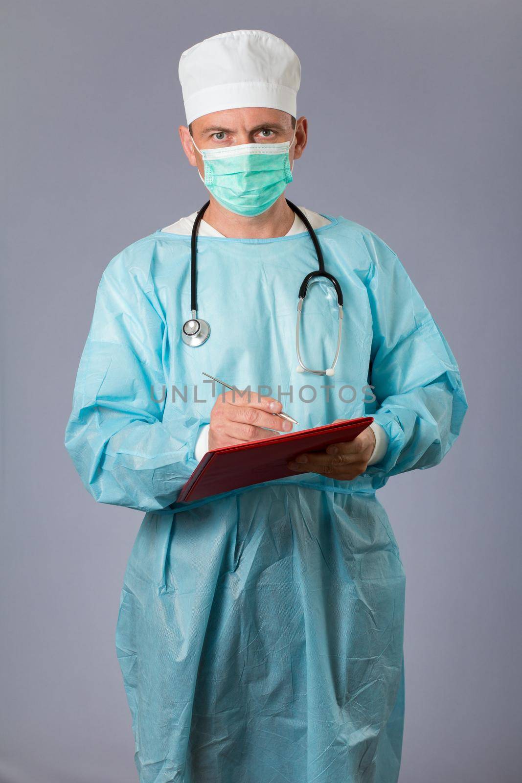 Medical doctor dressed in a medical gown with stethoscope and face mask holding a pen and writing in a notebook. Gray background