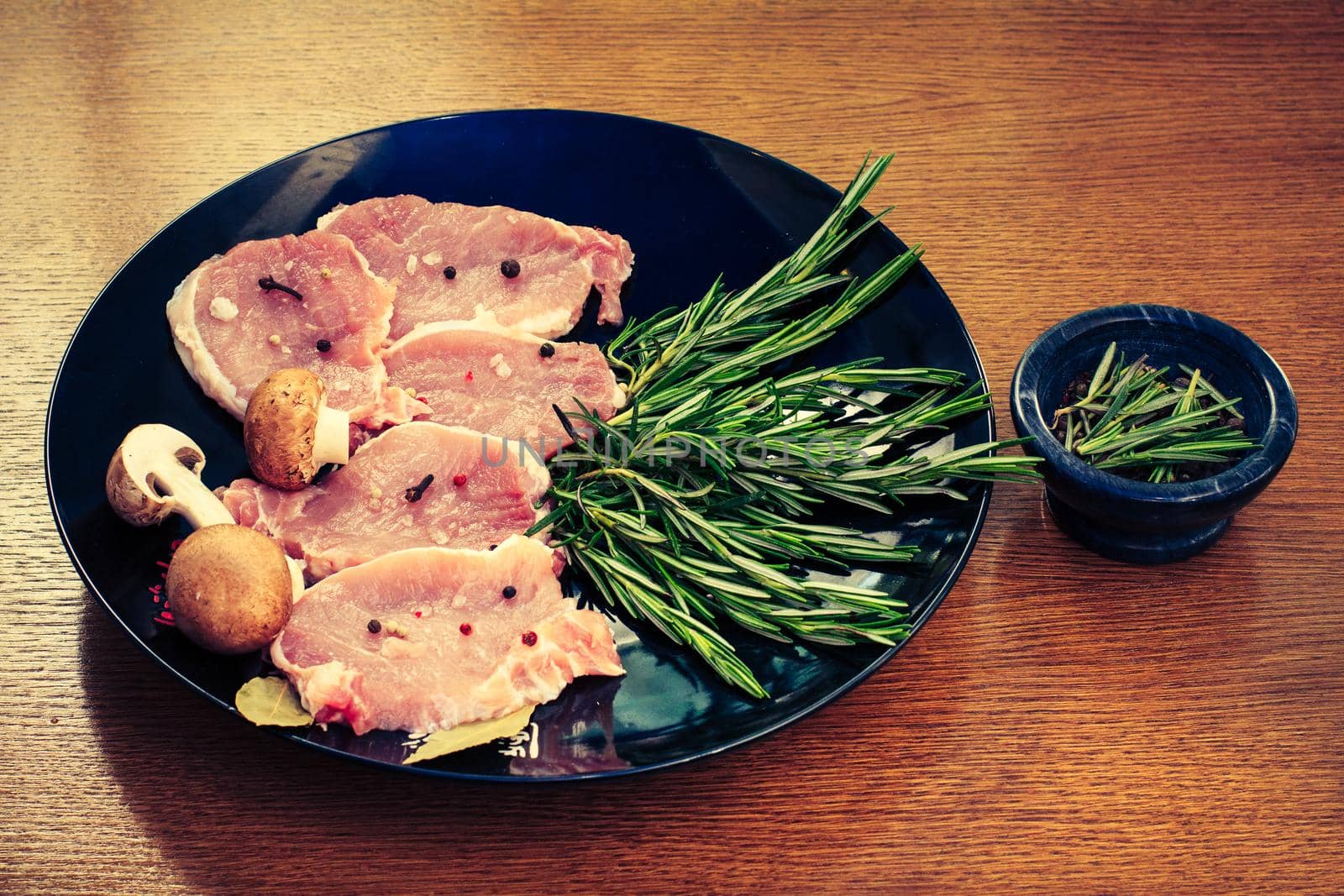 Pieces of raw pork steak with spices and herbs rosemary, mushrooms, salt and pepper on a black plate