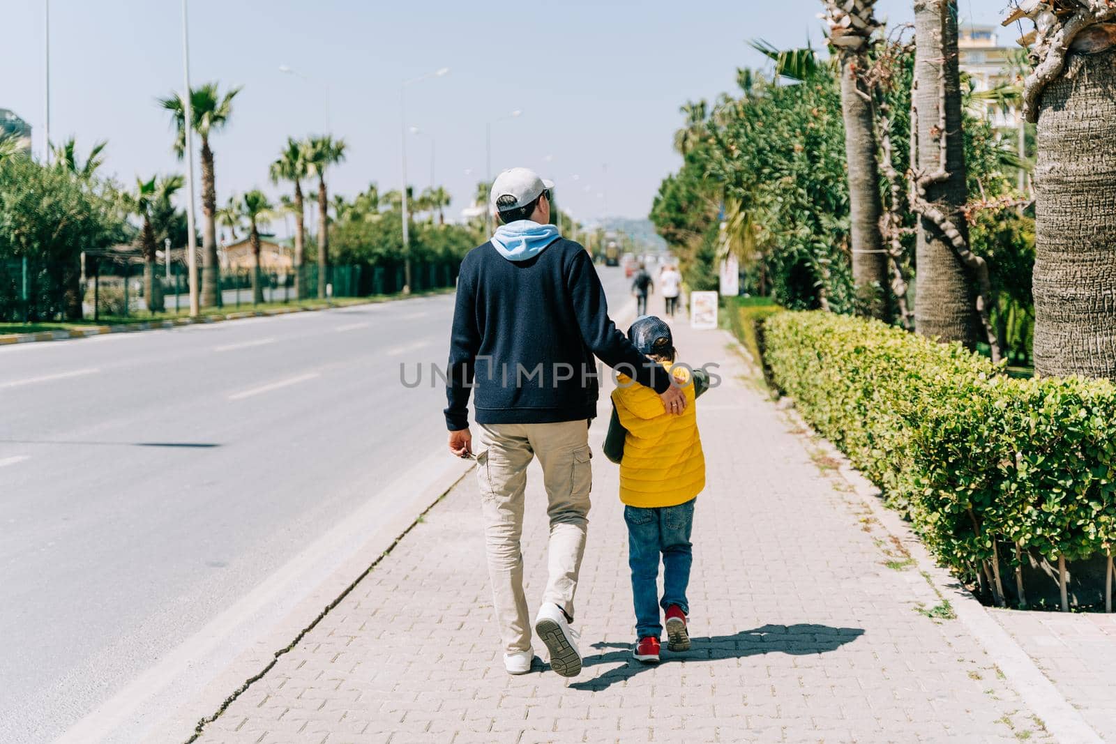 Rearview of father and son walking near city road. Dad holding his kid's back when taking a stroll in the southern city. Man and boy going for promenade near highway.