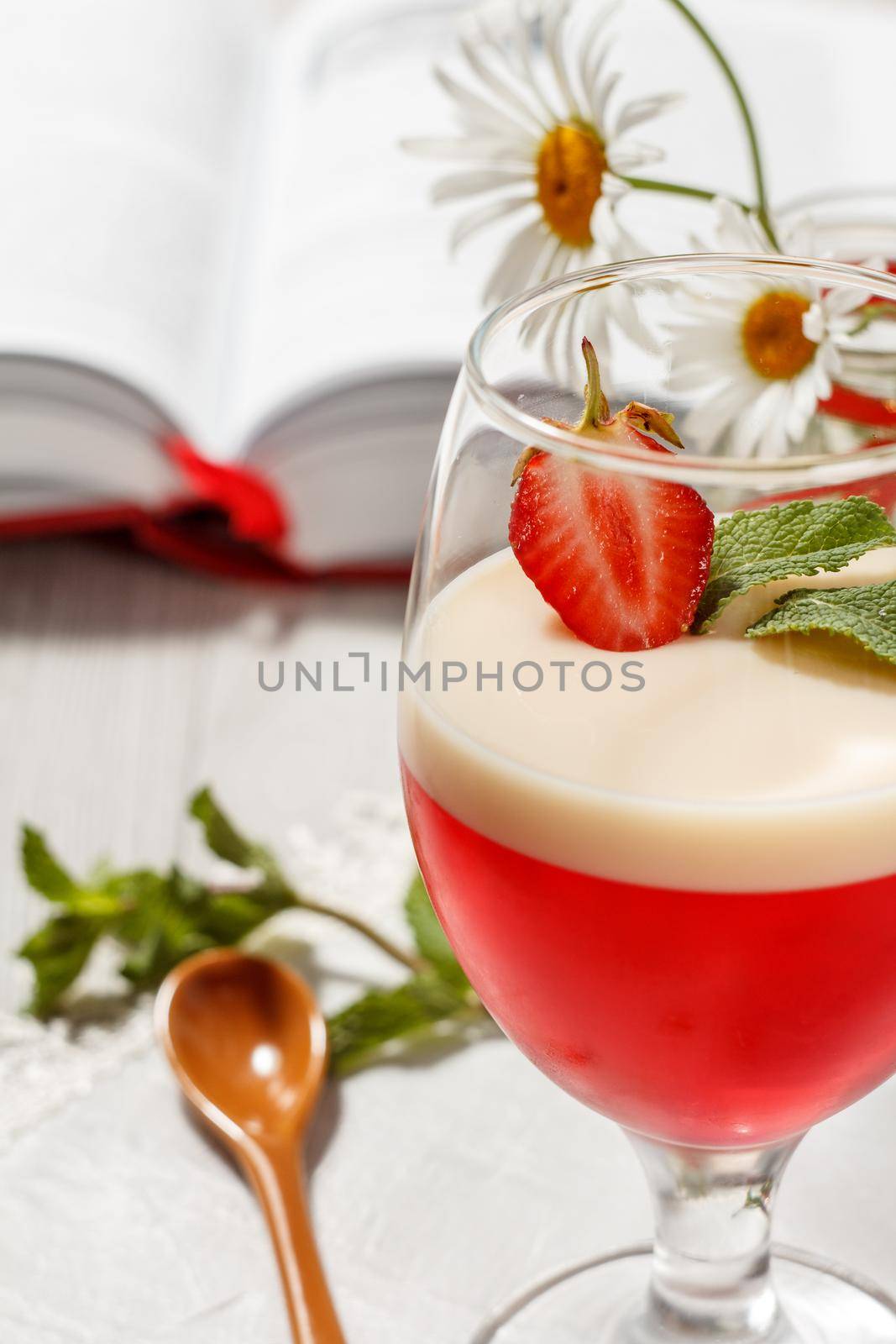 Cherry and milk jelly with strawberry piece in the glass topped mint leaves and opened book with chamomiles on the background