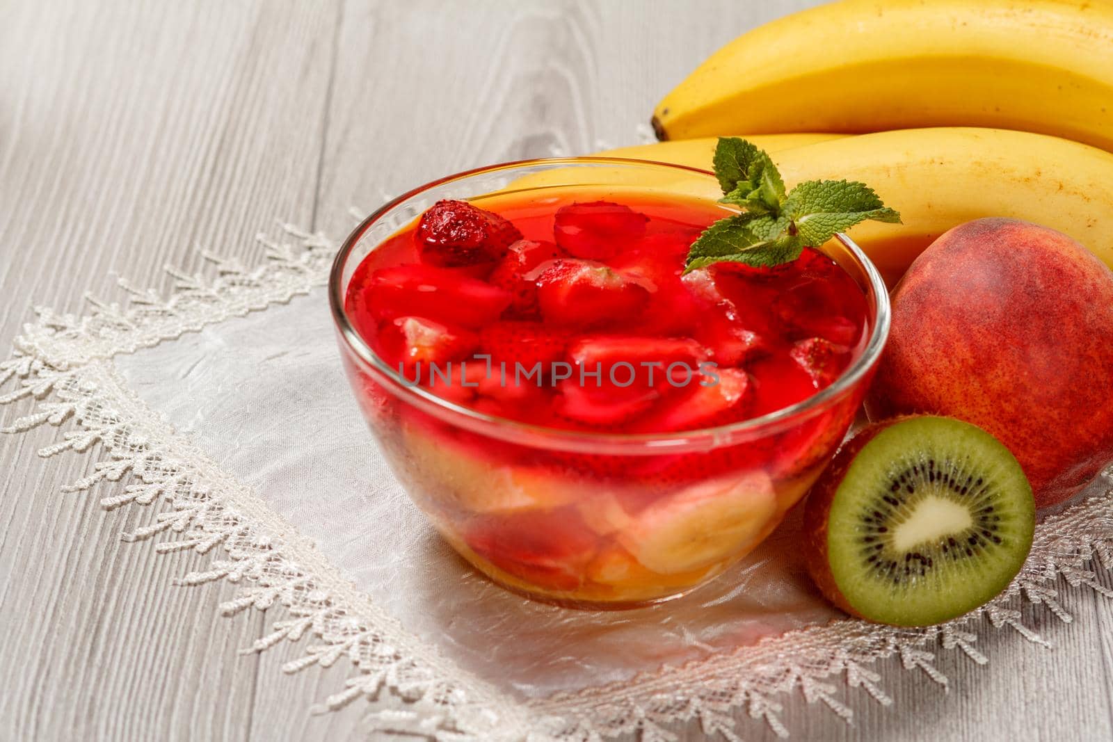 Cherry jelly with strawberry pieces in the glass bowl topped mint leaves, nectarine, kiwi and banana on the white napkin