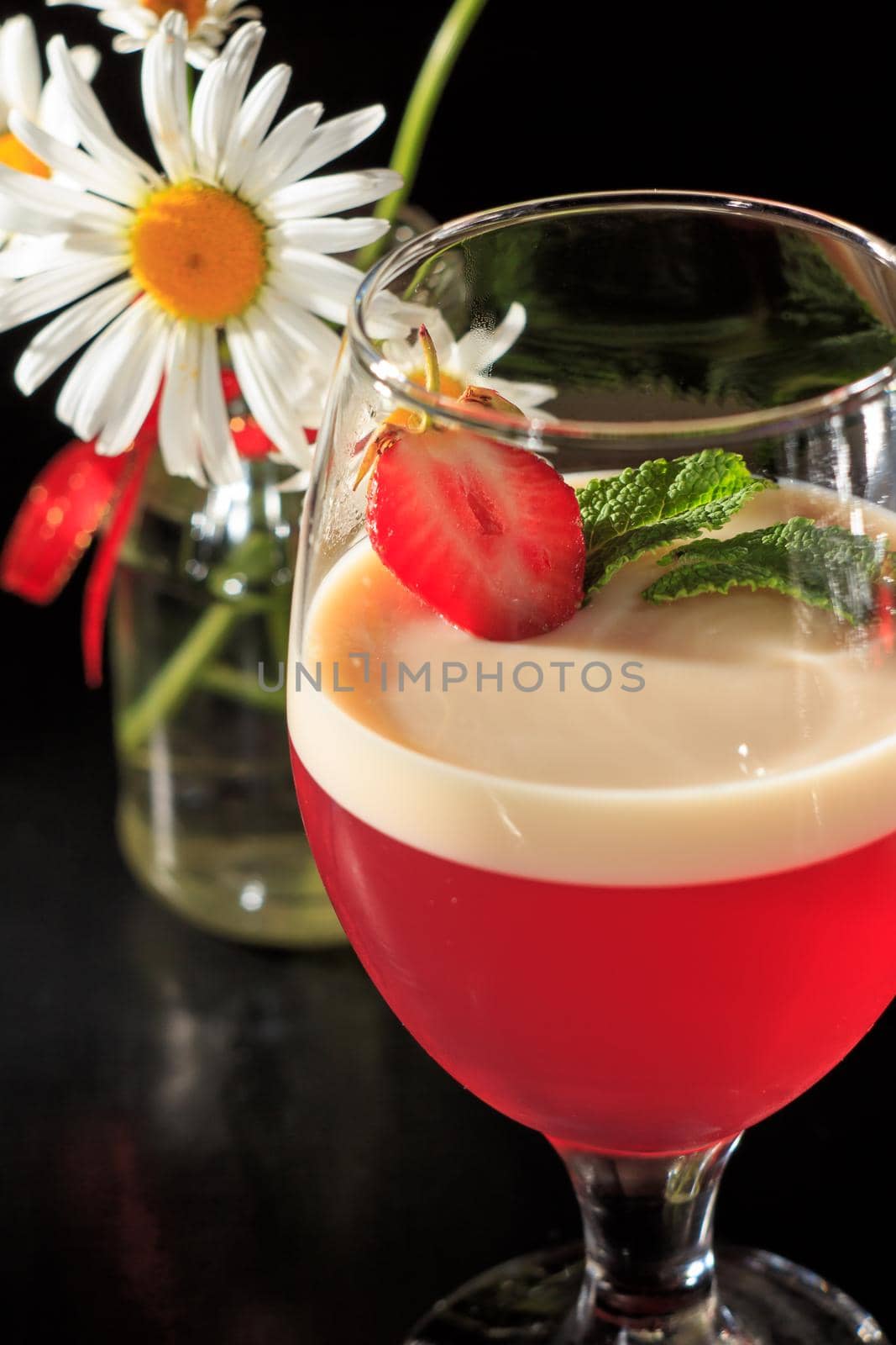 Cherry and milk jelly with strawberry piece and mint leaves in the glass with flower of chamomile on the black background