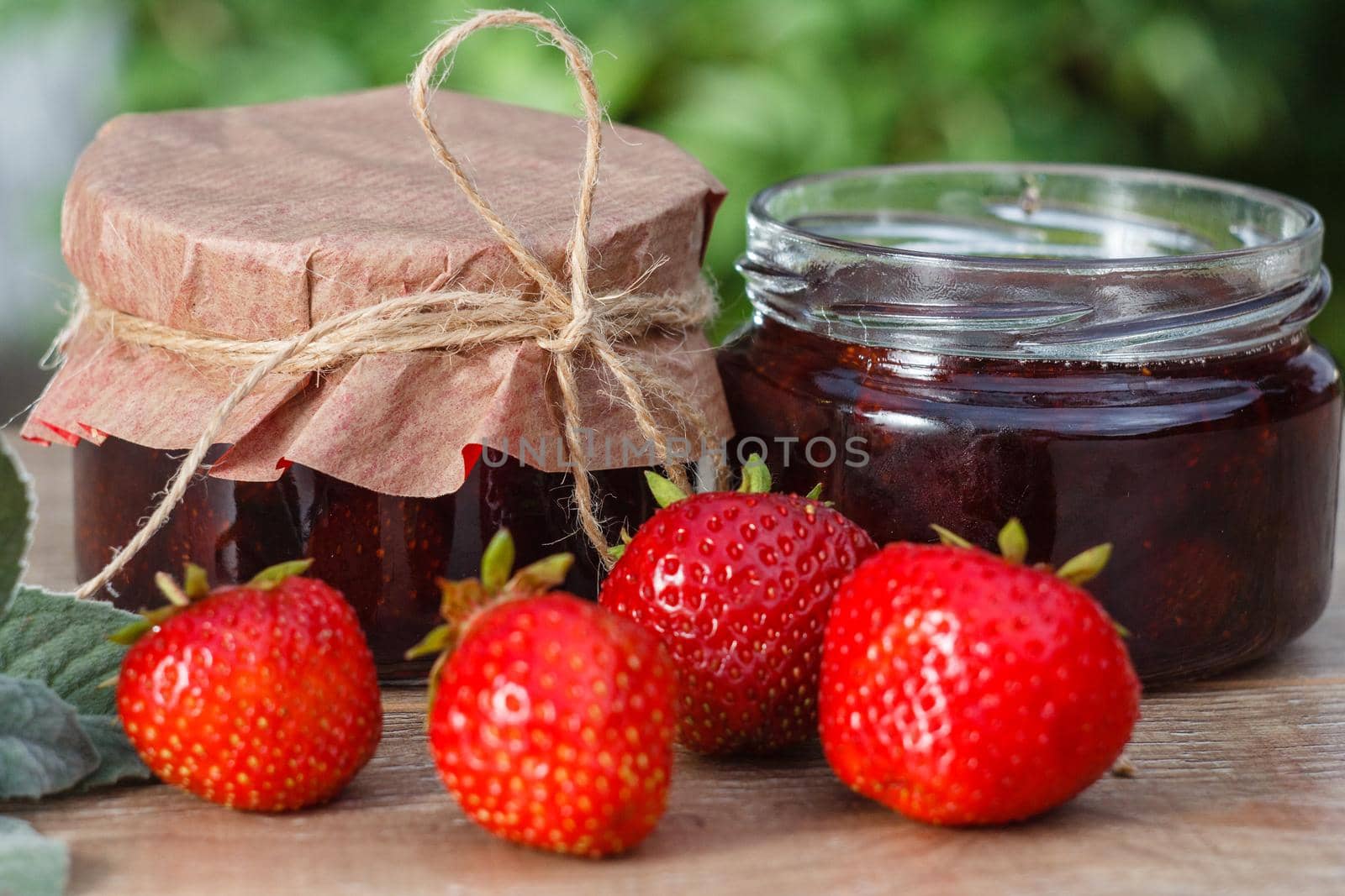 Traditional homemade strawberry jam in jars, decorated with fresh strawberries on wooden table with green blurred natural background Close up selective focus