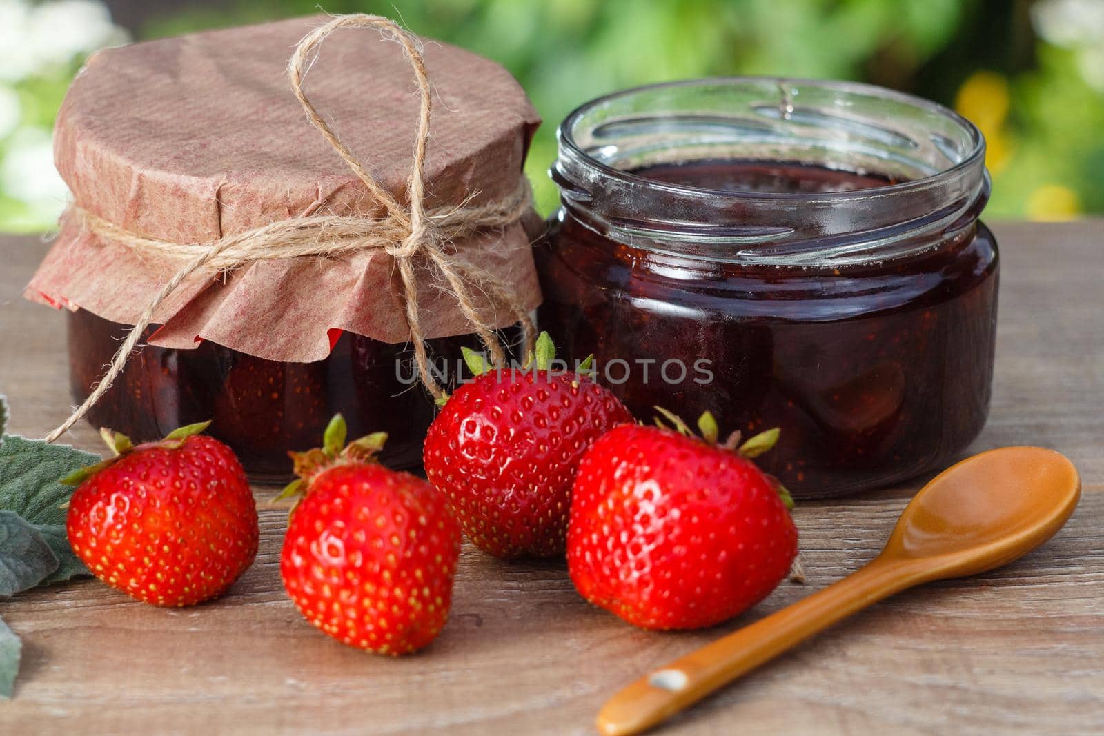 Traditional homemade strawberry jam in jars with spoon, decorated with fresh strawberries on wooden table with green blurred natural background Close up selective focus