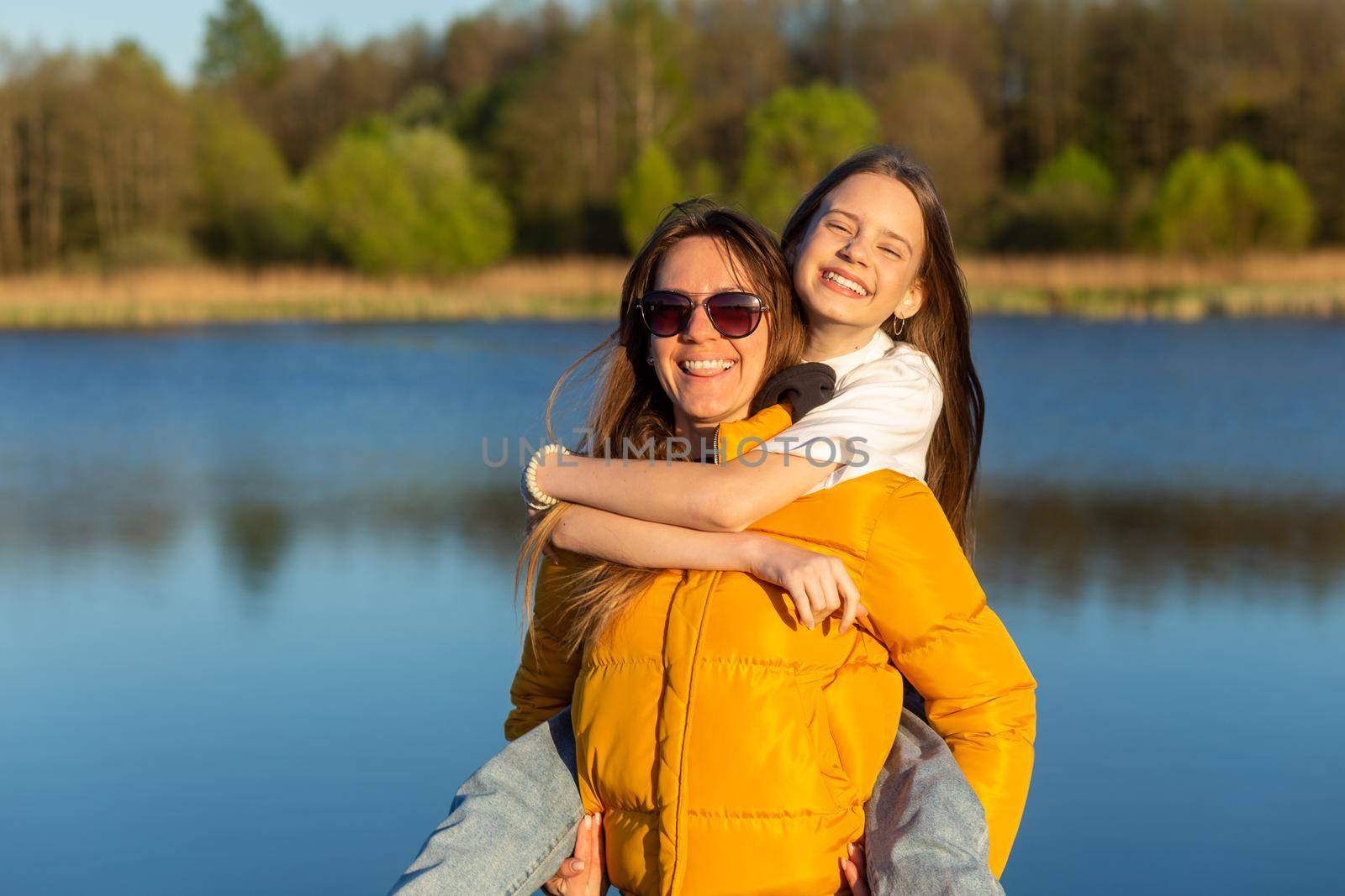 Playful mother giving daughter piggy back ride at spring lake shore. Both laughing and look happy. Spring in lake background. Closeup.