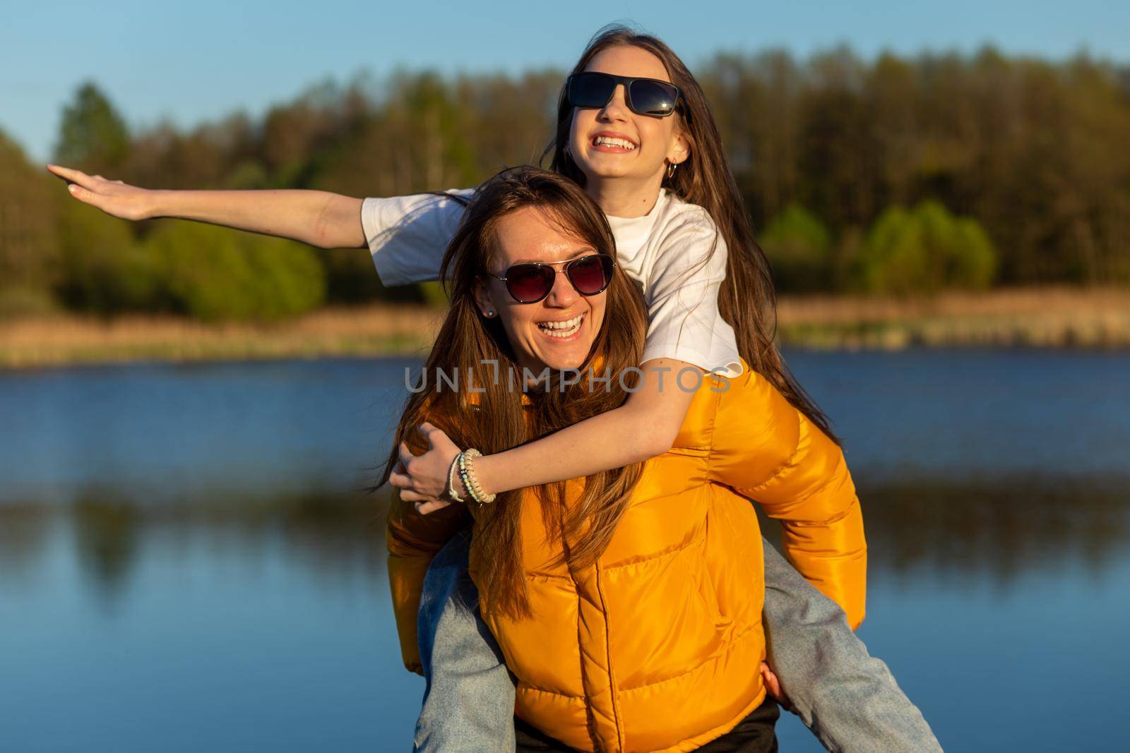 Playful mother giving daughter piggy back ride at spring lake shore. Both laughing and look happy. Spring in lake background. Closeup.