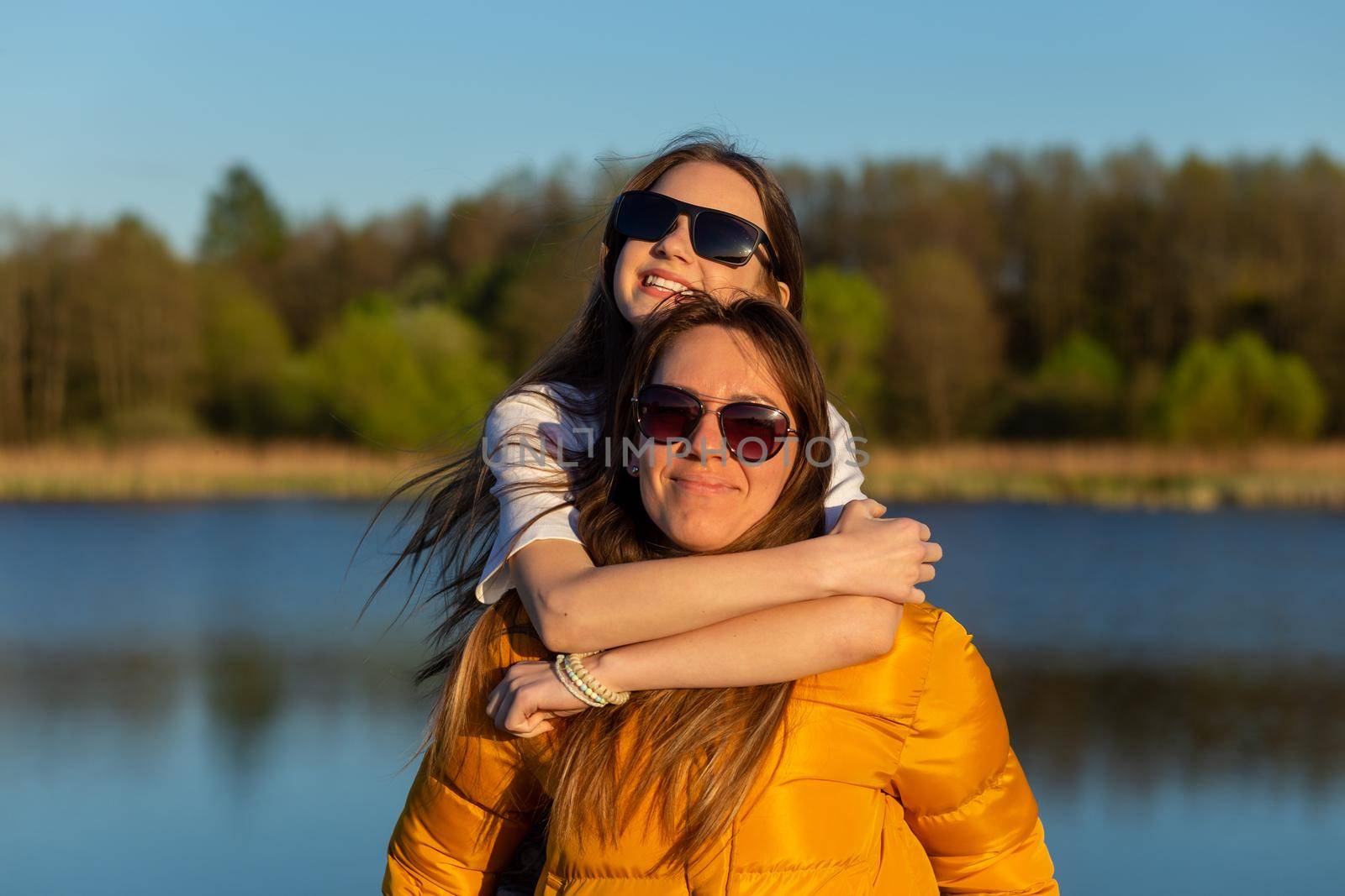 Playful mother giving daughter piggy back ride at spring lake shore. Both laughing and look happy. Spring in lake background. Closeup.