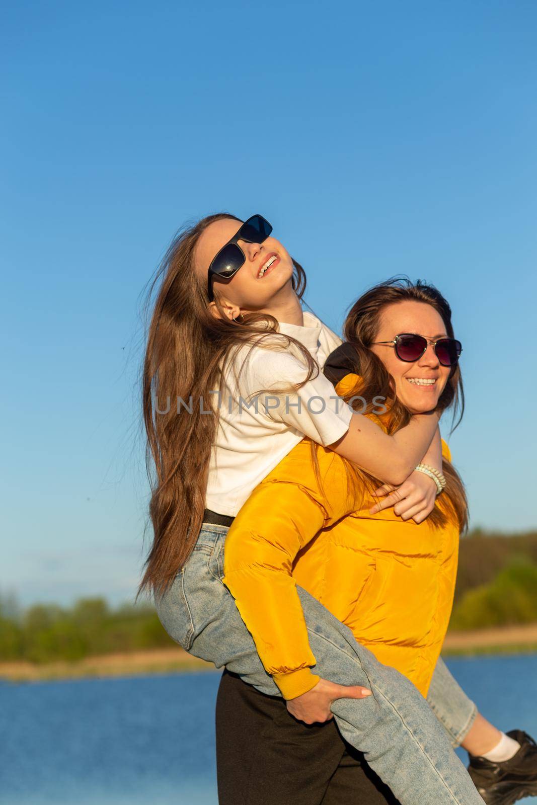 Playful mother giving daughter piggy back ride at spring lake shore. Both laughing and look happy. Spring in lake background. Closeup.