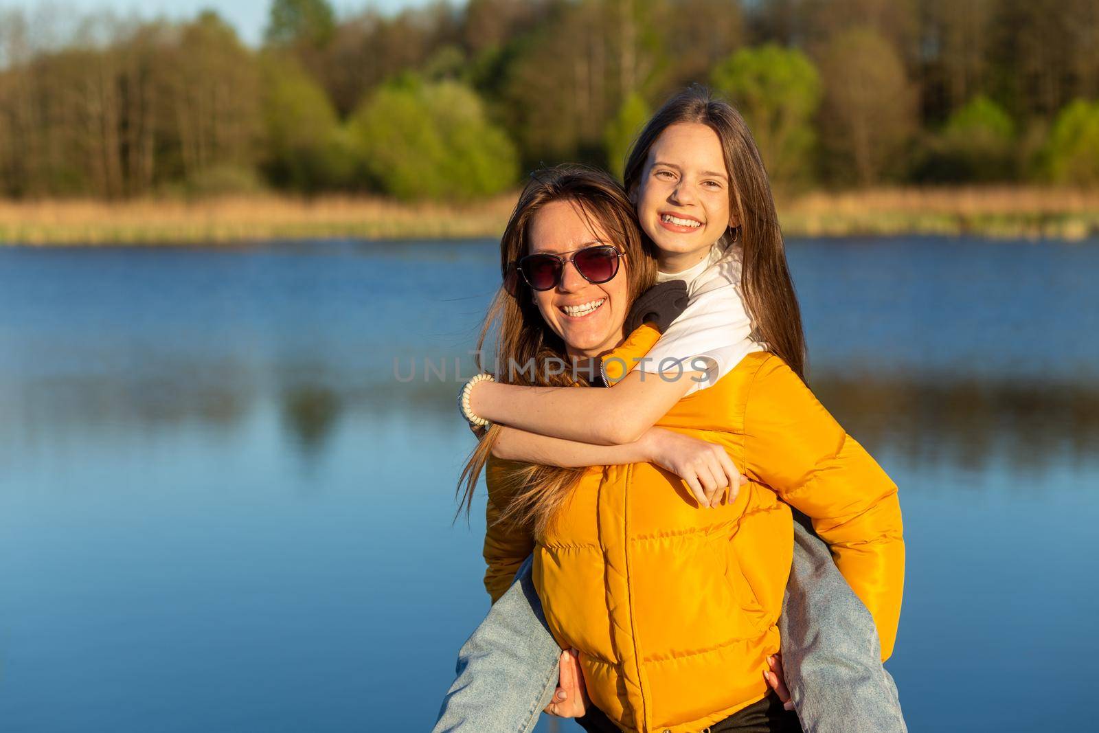 Playful mother giving daughter piggy back ride at spring lake shore. by BY-_-BY