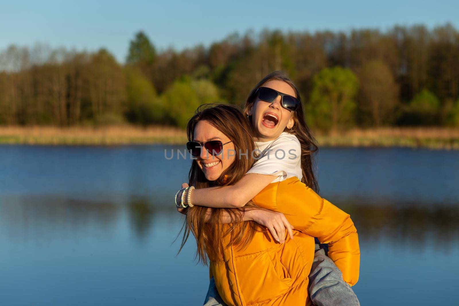 Playful mother giving daughter piggy back ride at spring lake shore. Both laughing and look happy. Spring in lake background. Closeup.