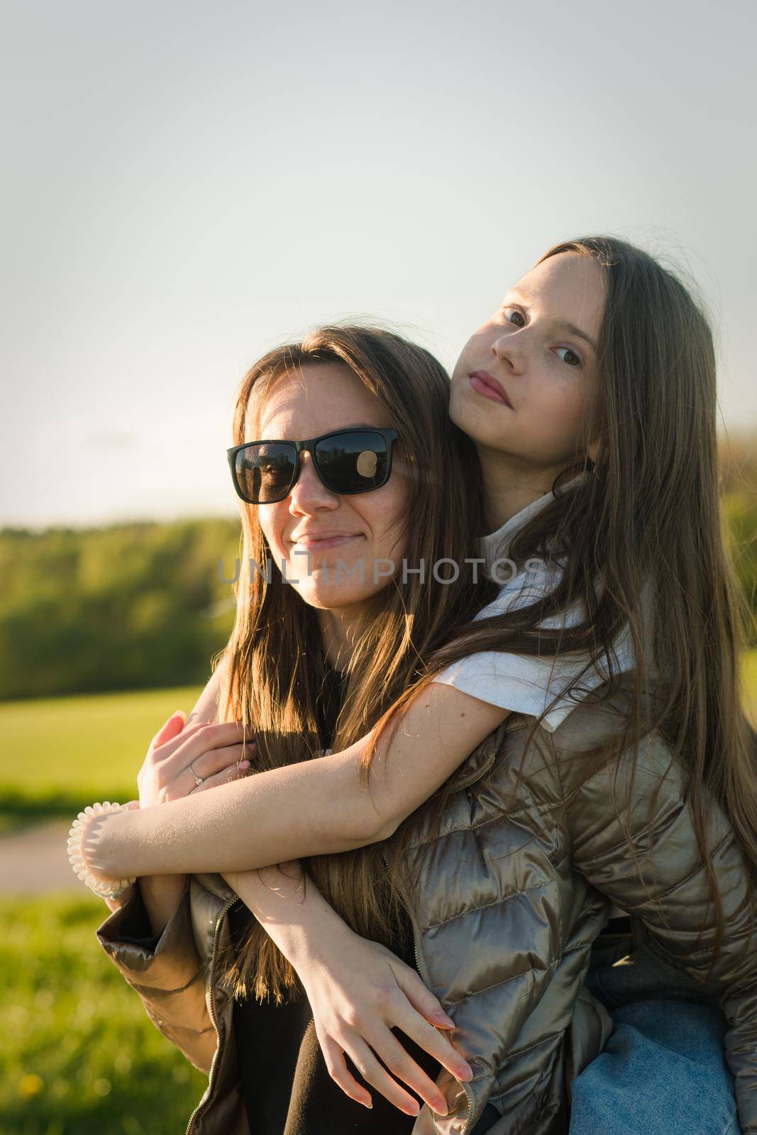 Playful mother giving daughter piggy back ride at green field. Both laughing and look happy. Spring in forest background. Closeup.