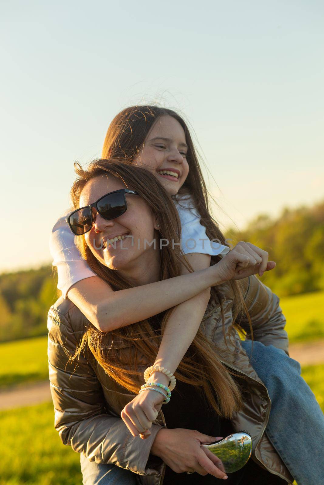 Playful mother giving daughter piggy back ride at green field. Both laughing and look happy. Spring in forest background. Closeup.