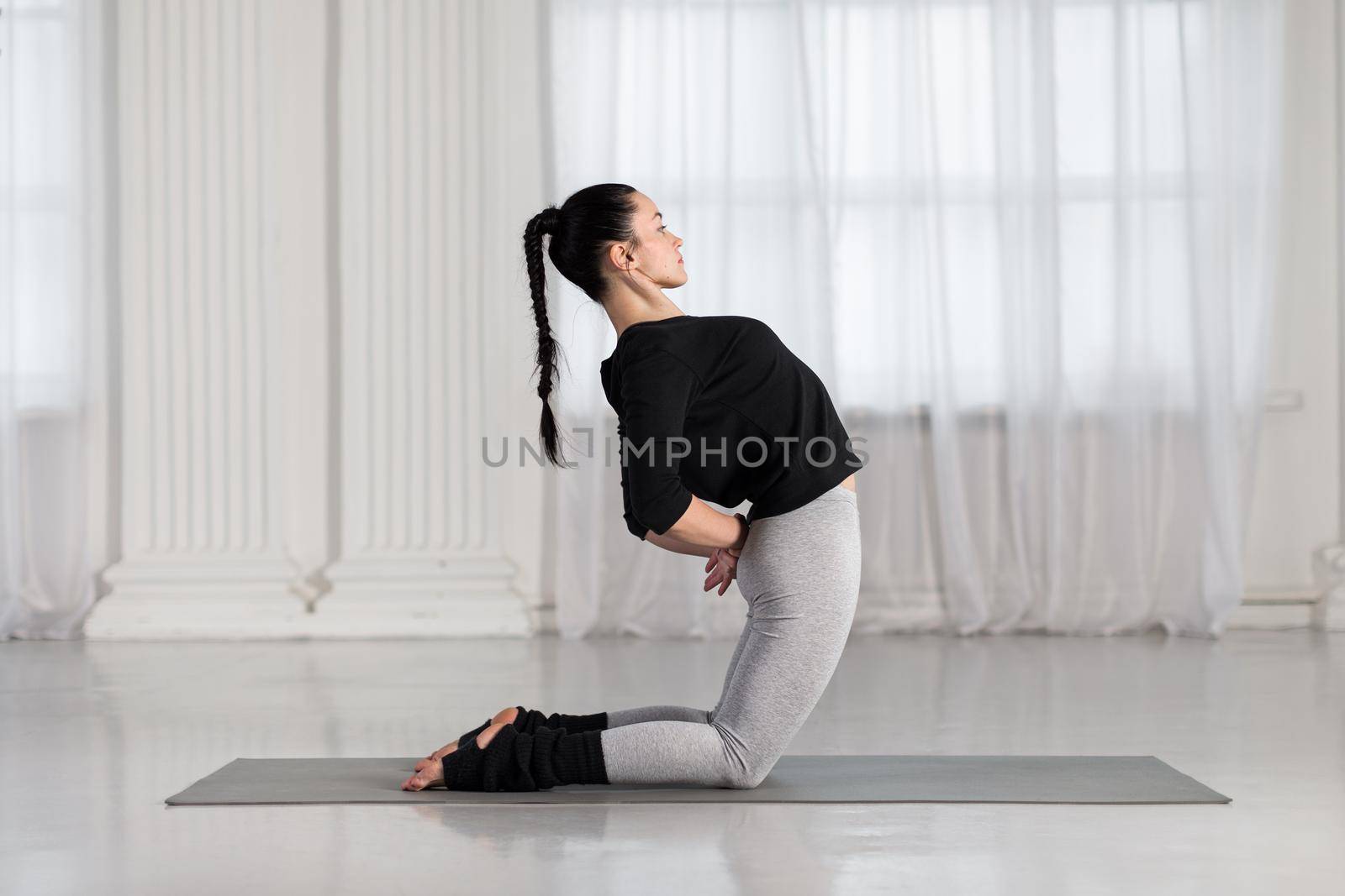 Beautiful young woman working out indoors, doing yoga exercise on gray mat, stretching and preparing for standing in Ustrasana, Camel Posture, full length, copy space