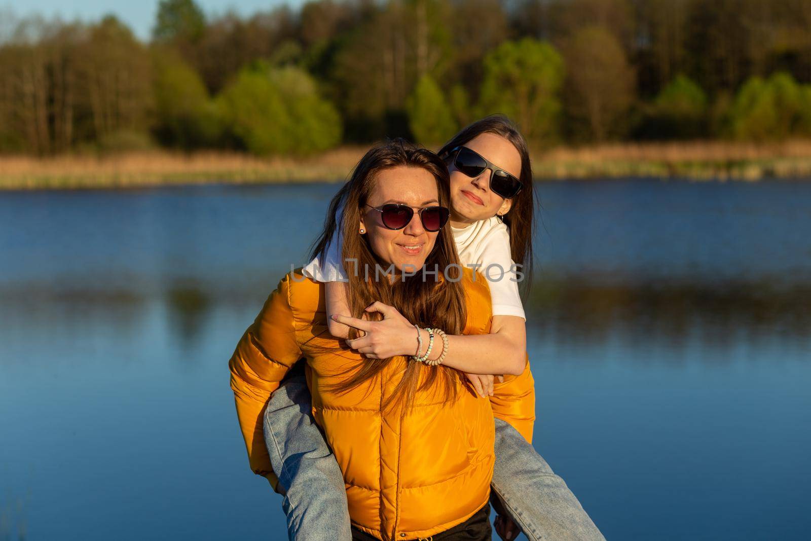 Playful mother giving daughter piggy back ride at spring lake shore. Both laughing and look happy. Spring in lake background. Closeup.