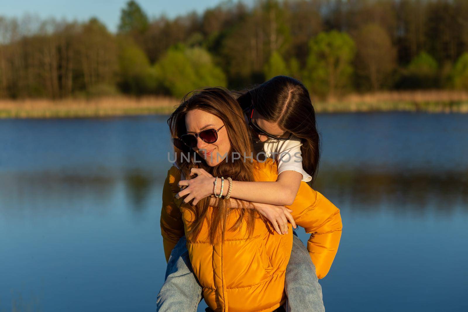 Playful mother giving daughter piggy back ride at spring lake shore. Both laughing and look happy. Spring in lake background. Closeup.