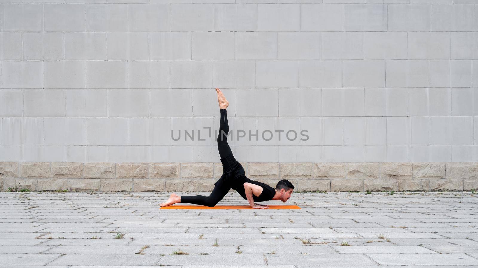 Chaturanga pose with one leg up. Fit latin man do yoga outdoor on orange mat with gray concrete wall at the background. Copy space.