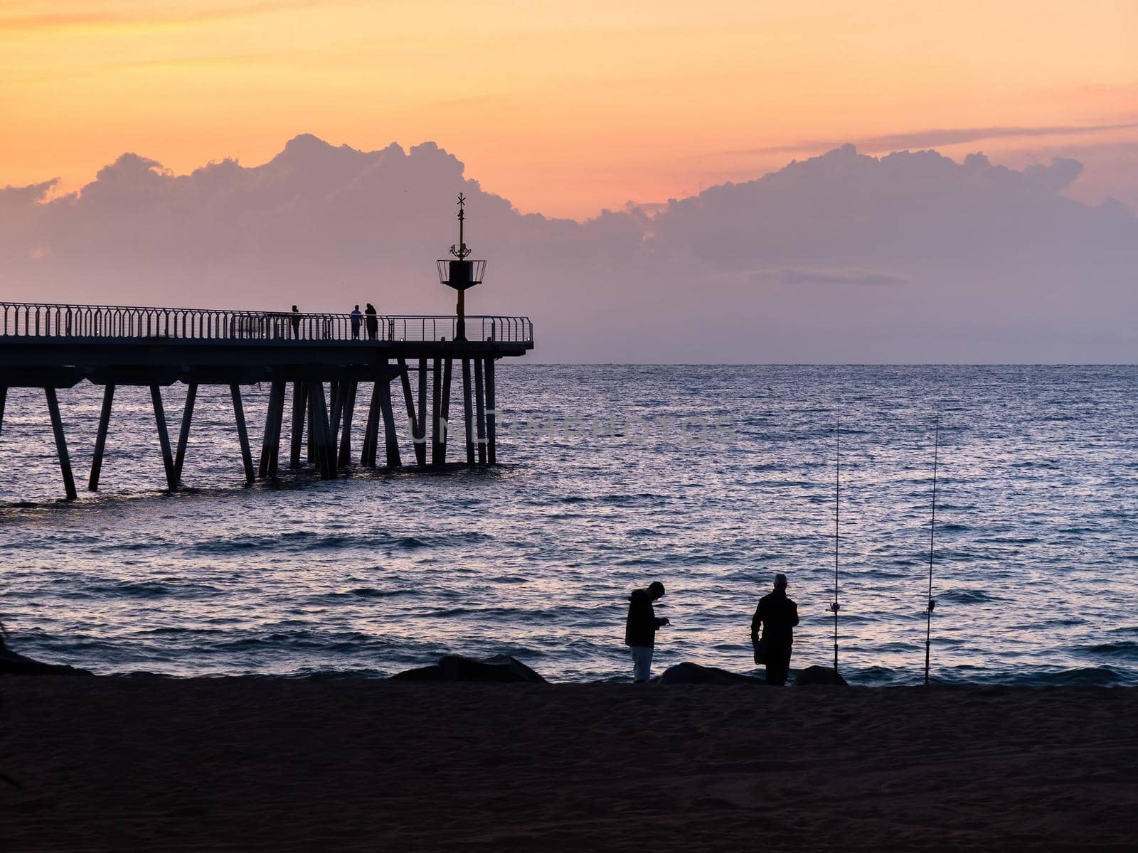 Two fishermen are fishing on beach at the sunset using fishing rods, set against the coast. Pontoon, orange sky and clouds at the background. Badalona, Barcelona, Spain