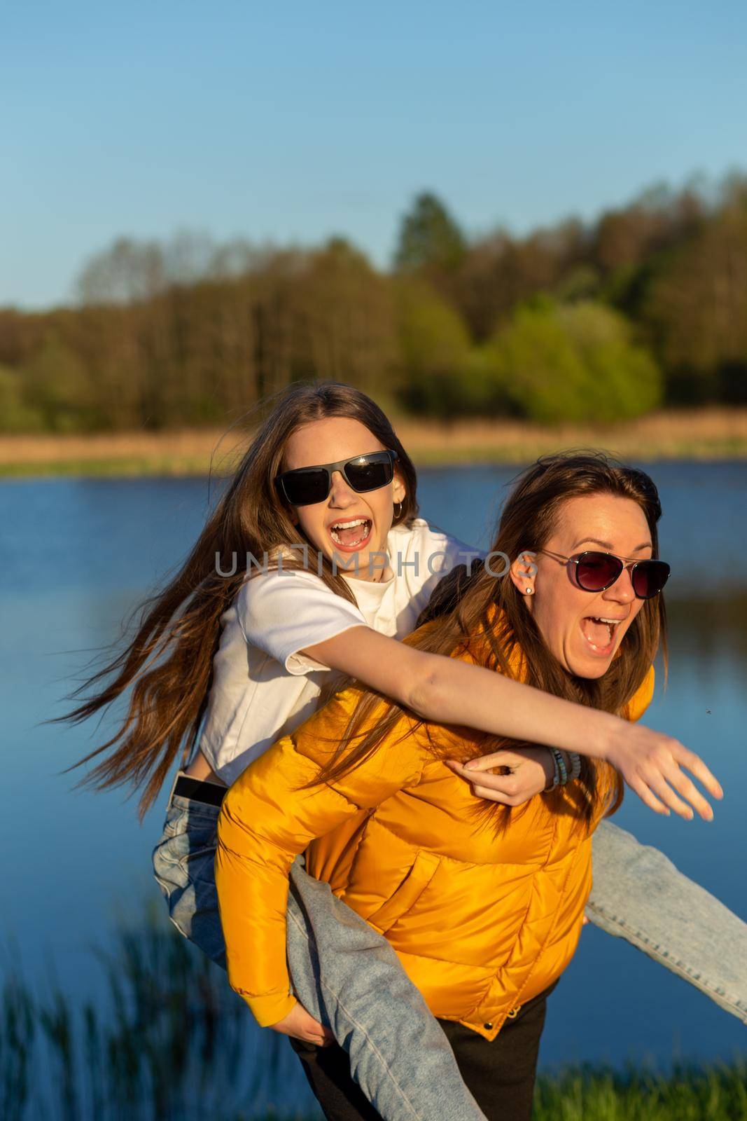 Playful mother giving daughter piggy back ride at spring lake shore. by BY-_-BY
