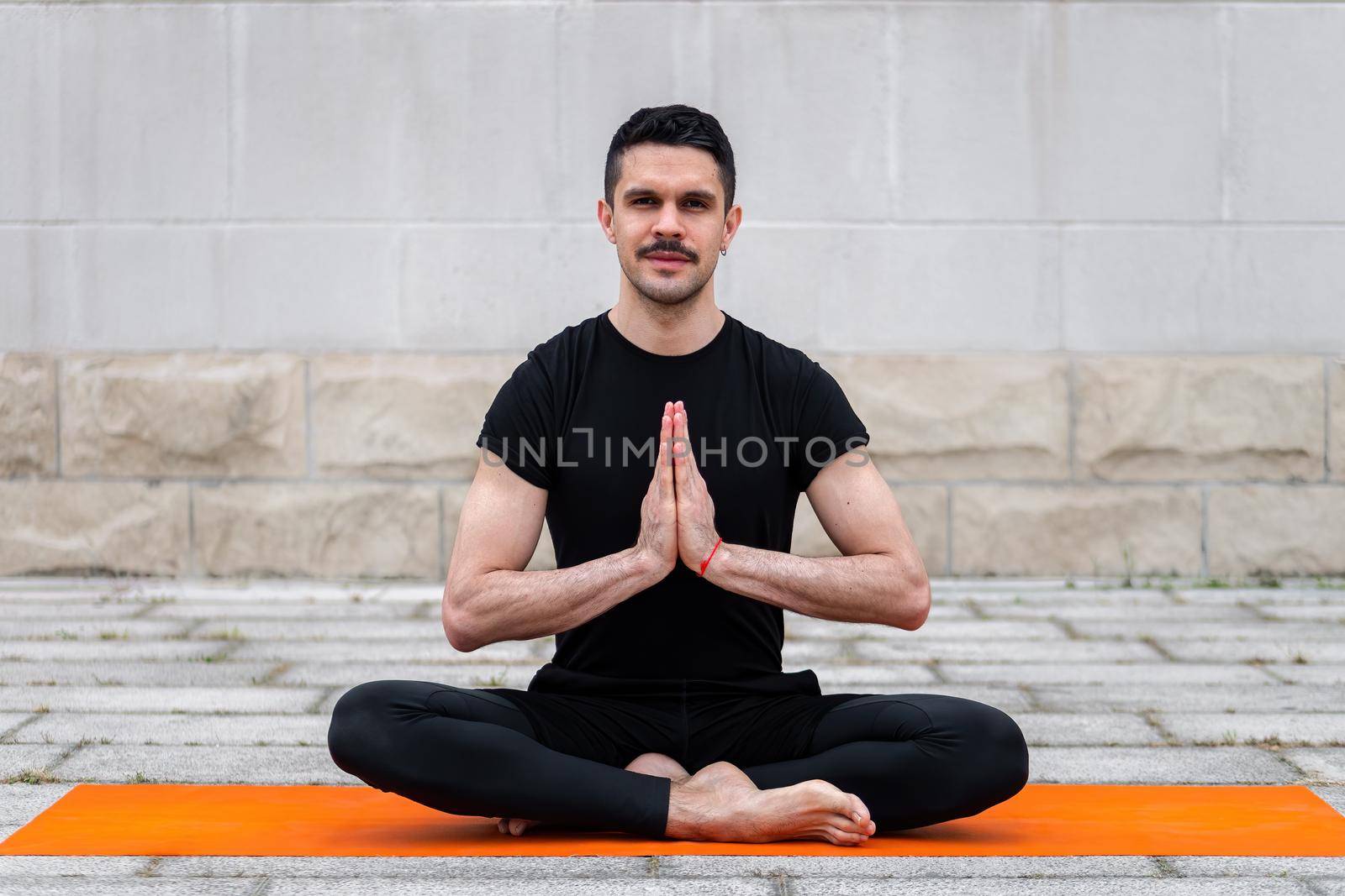 Latin gay practicing yoga outdoor in a city, sitting in yoga pose on orange mat, with gray wall at the background. by apavlin