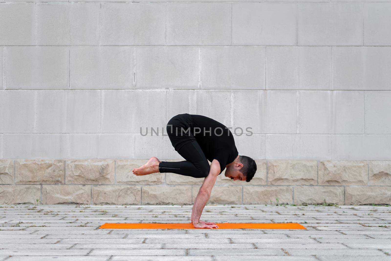 Latin man practicing yoga in a city, standing in crane pose on orange mat, with gray wall at the background. Side view with copy space