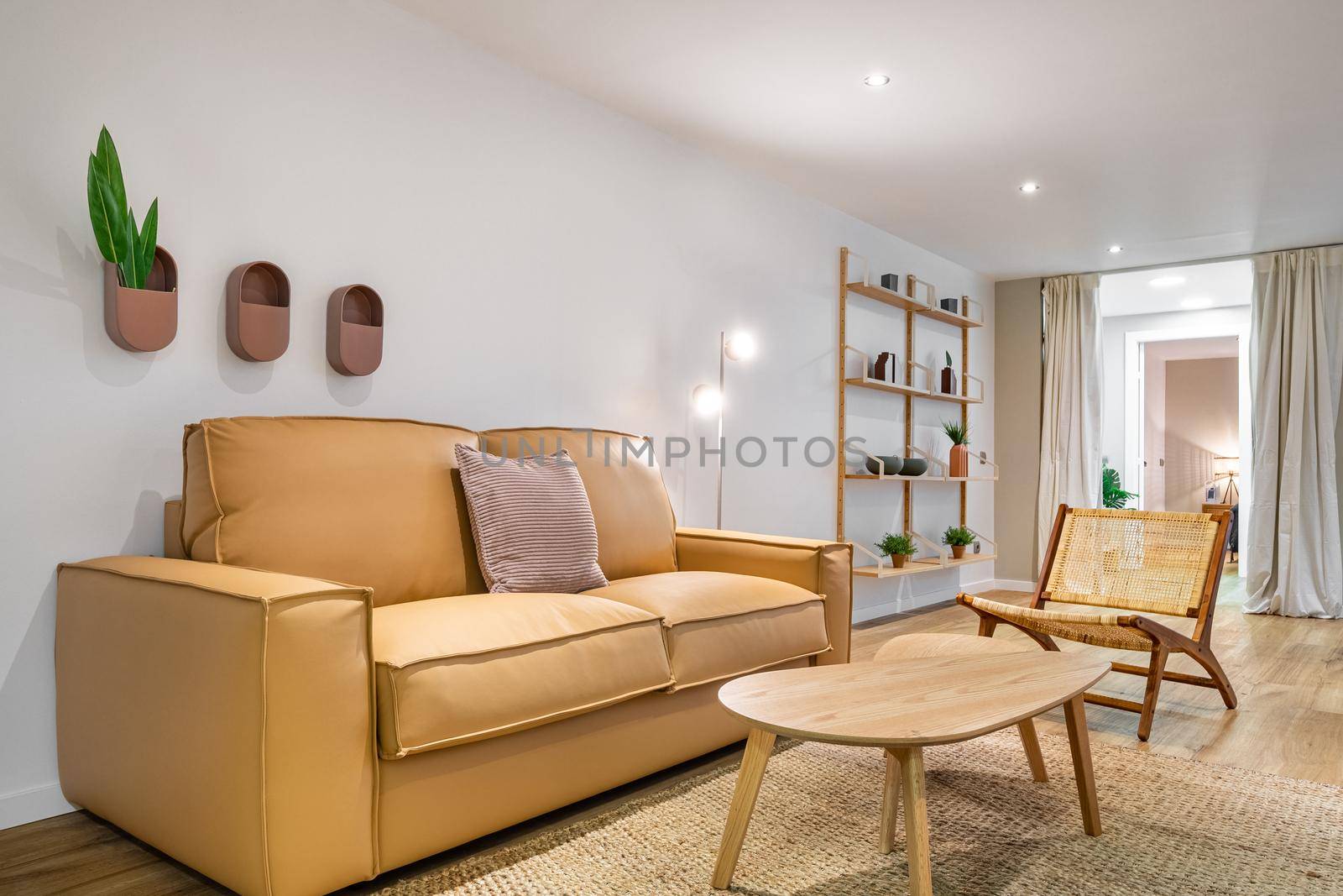 Modern boho interior of living room at cozy apartment with beige sofa, wooden table, wicker chair and stand with shelves at the background. Stylish home decor.