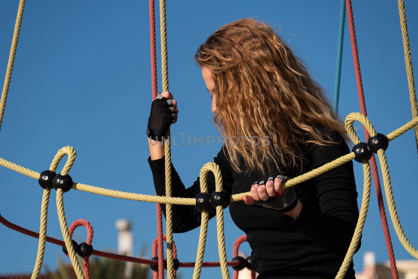 Active curly girl in black clothes playing on climbing net at playground in the park. Sport outside on sunny summer day. Young sportive woman having fun at recreational center.