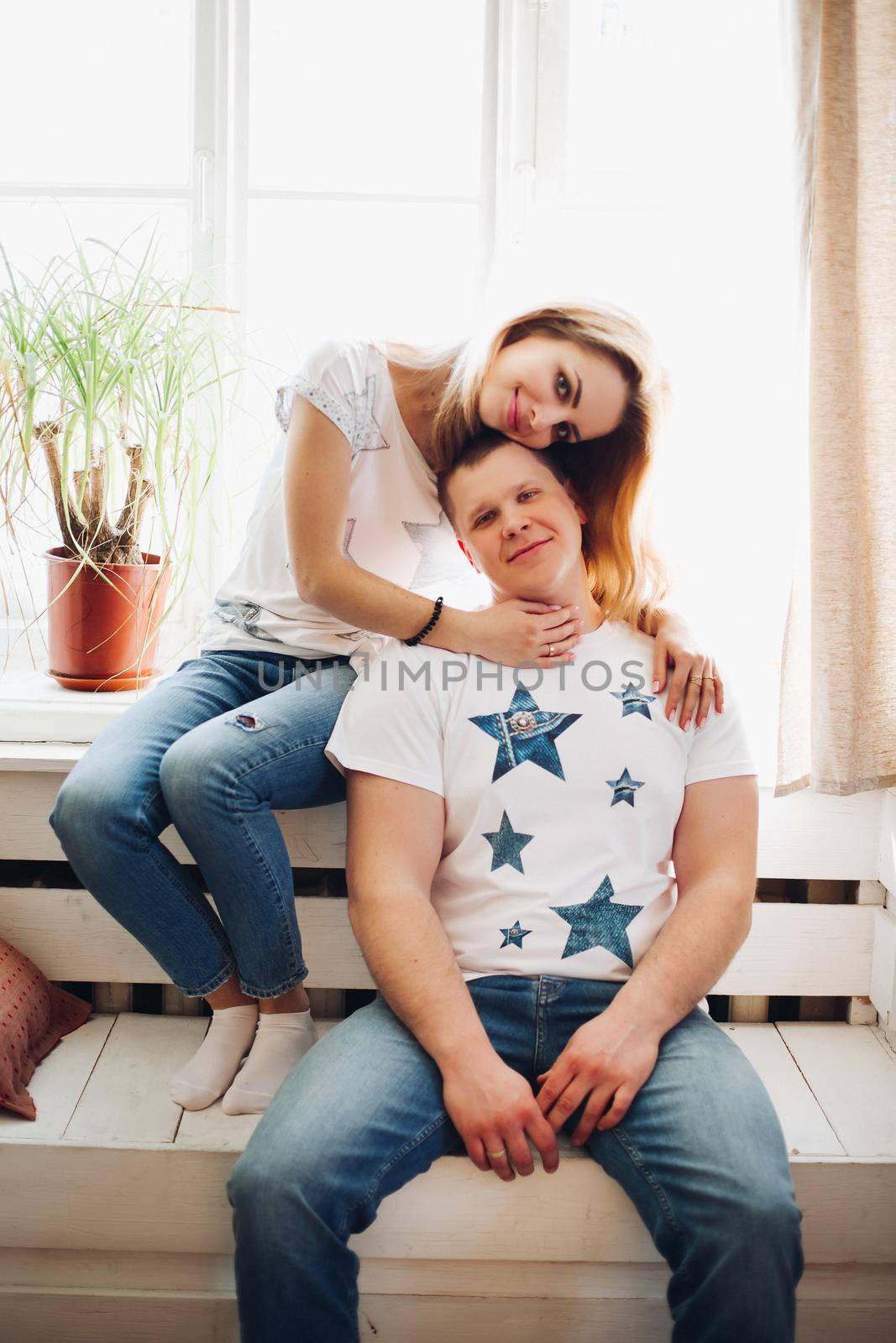 Portrait of romantic couple, sitting on white wooden bench in front of large window. Boyfriend and girlfriend wearing in white t-shirt and jeans embracing and looking at camera. Concept of love.