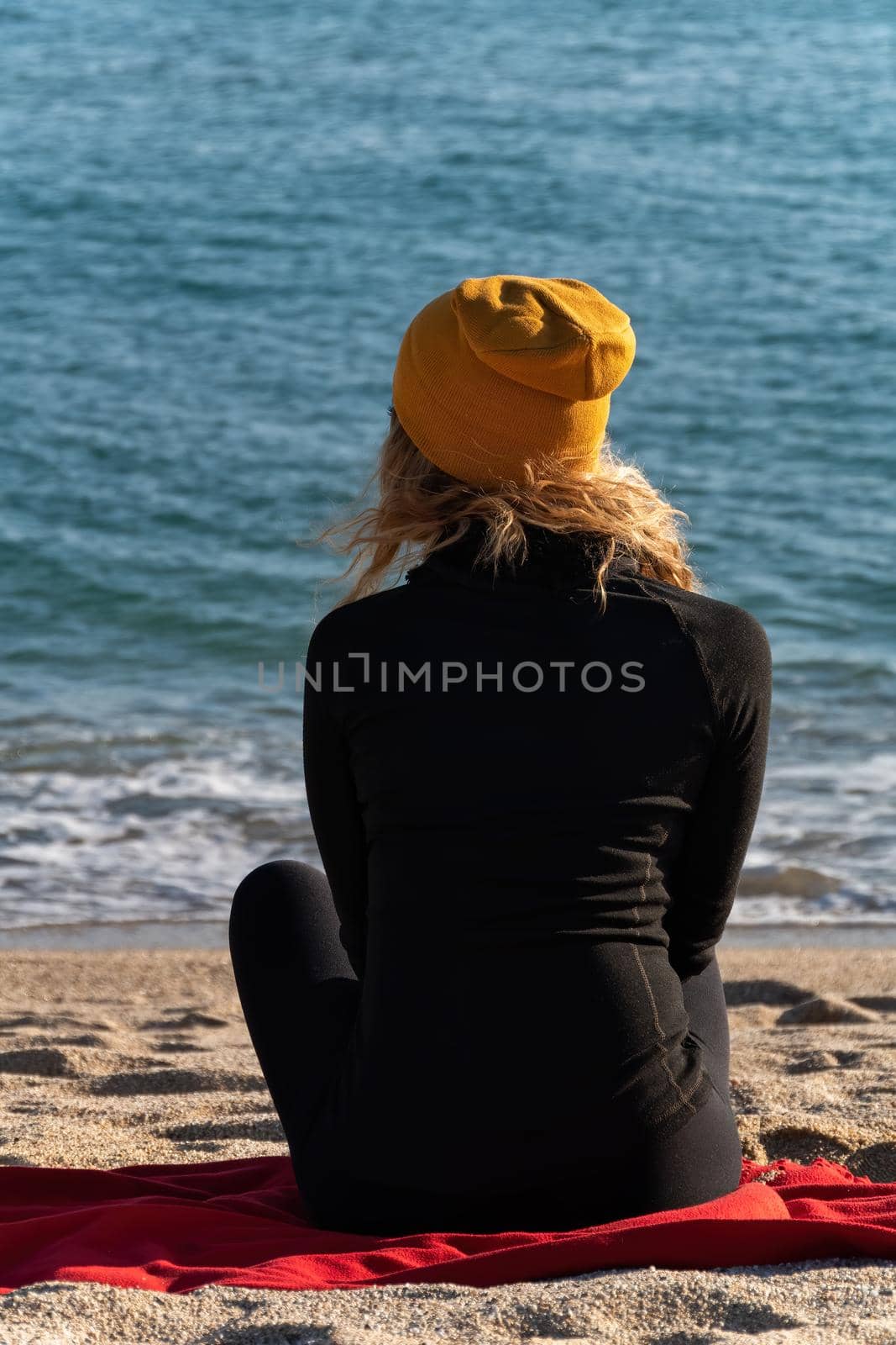A girl with blond curly hair in black sport jacket and yellow cap is sitting on the beach and looking at the blue sea. View from the back.