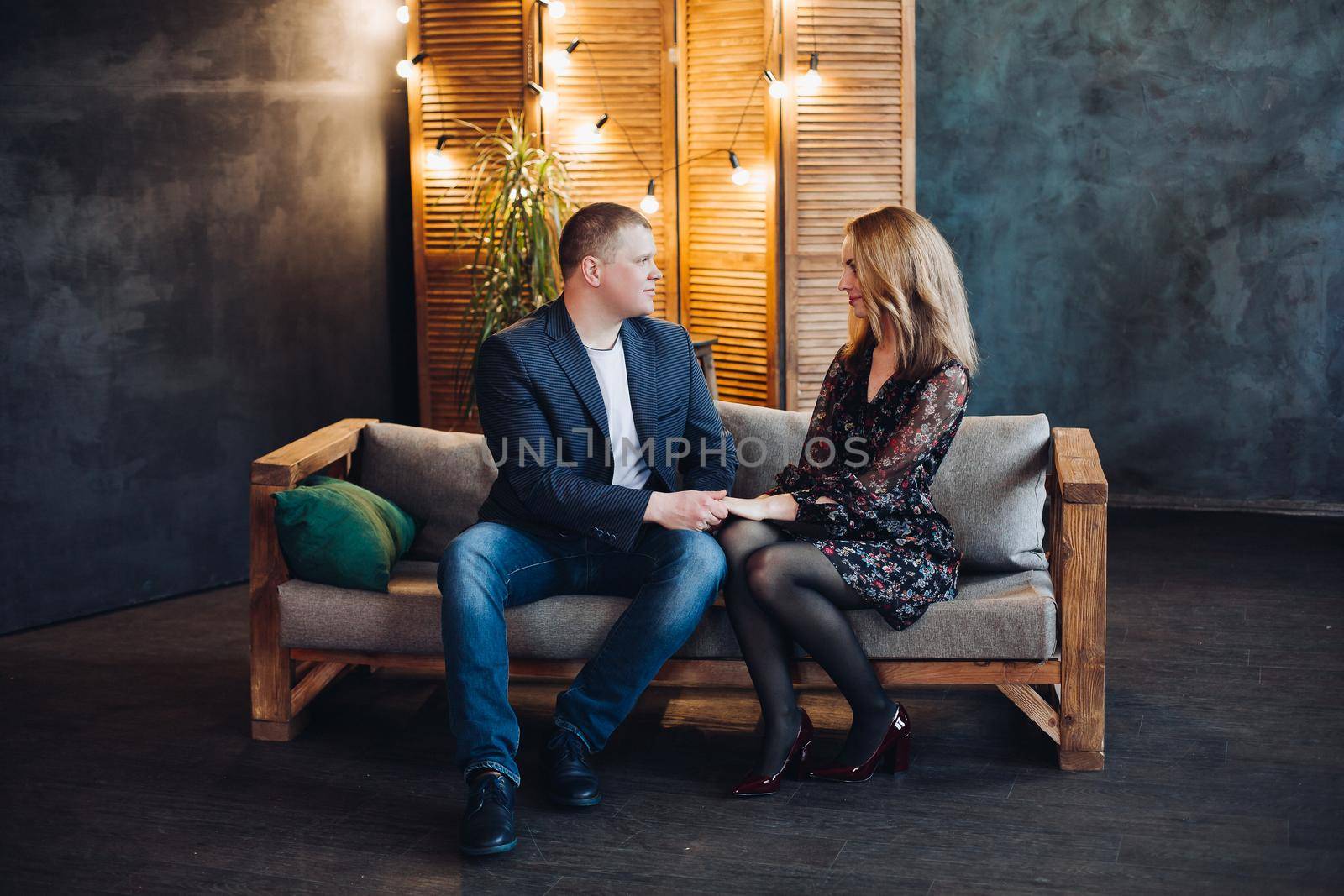 Portrait of romantic couple in love, looking in eyes of each other, sitting on grey couch in decorated studio. Boyfriend tenderly holding hand of his beautiful blonde girlfriend in black dress.