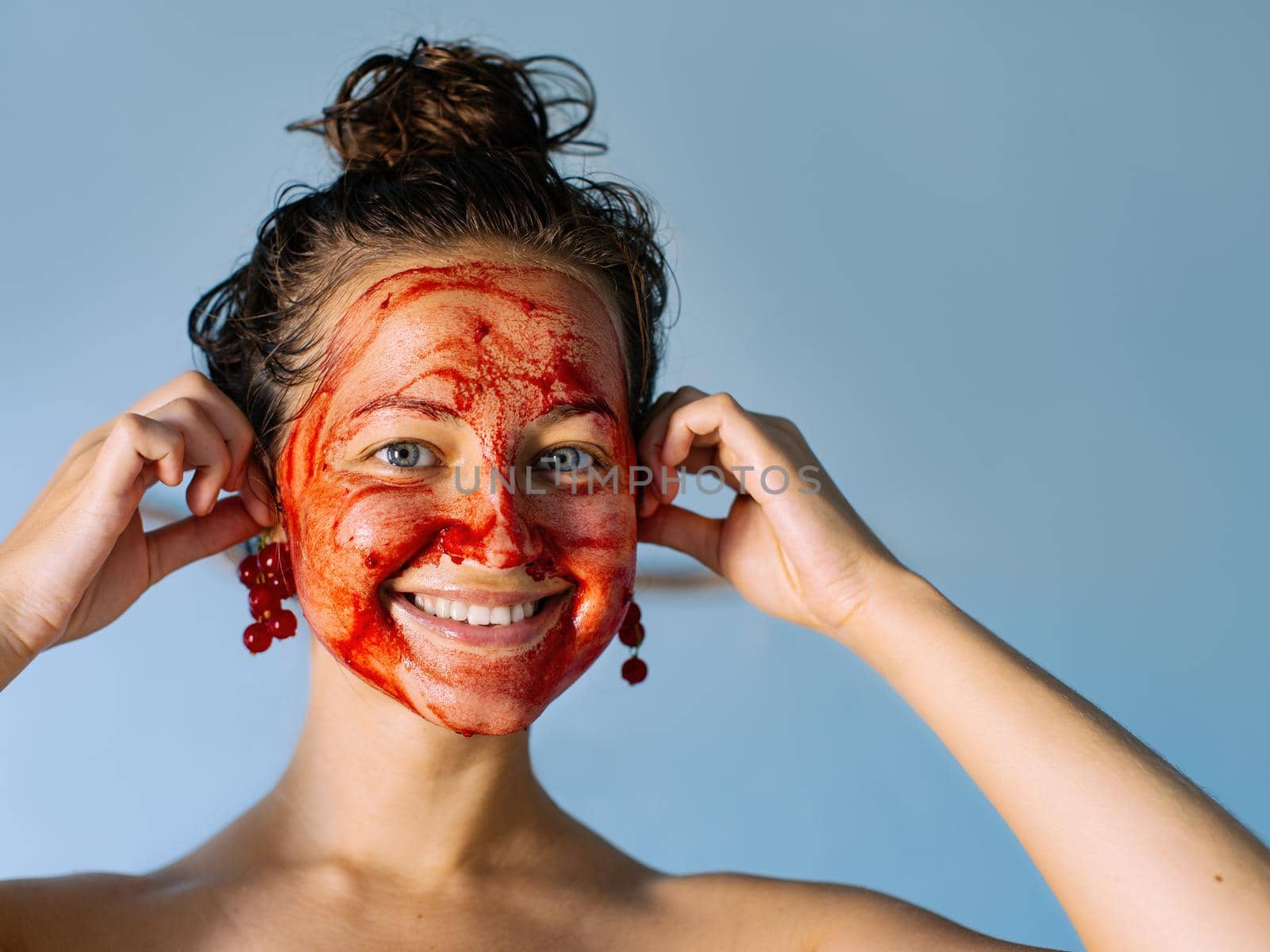 Beautiful young girl with a refreshing fruit mask from redcurrant on her face, smiles and holds twigs of red currant like earrings. Beauty and skin care at home. Copy space.