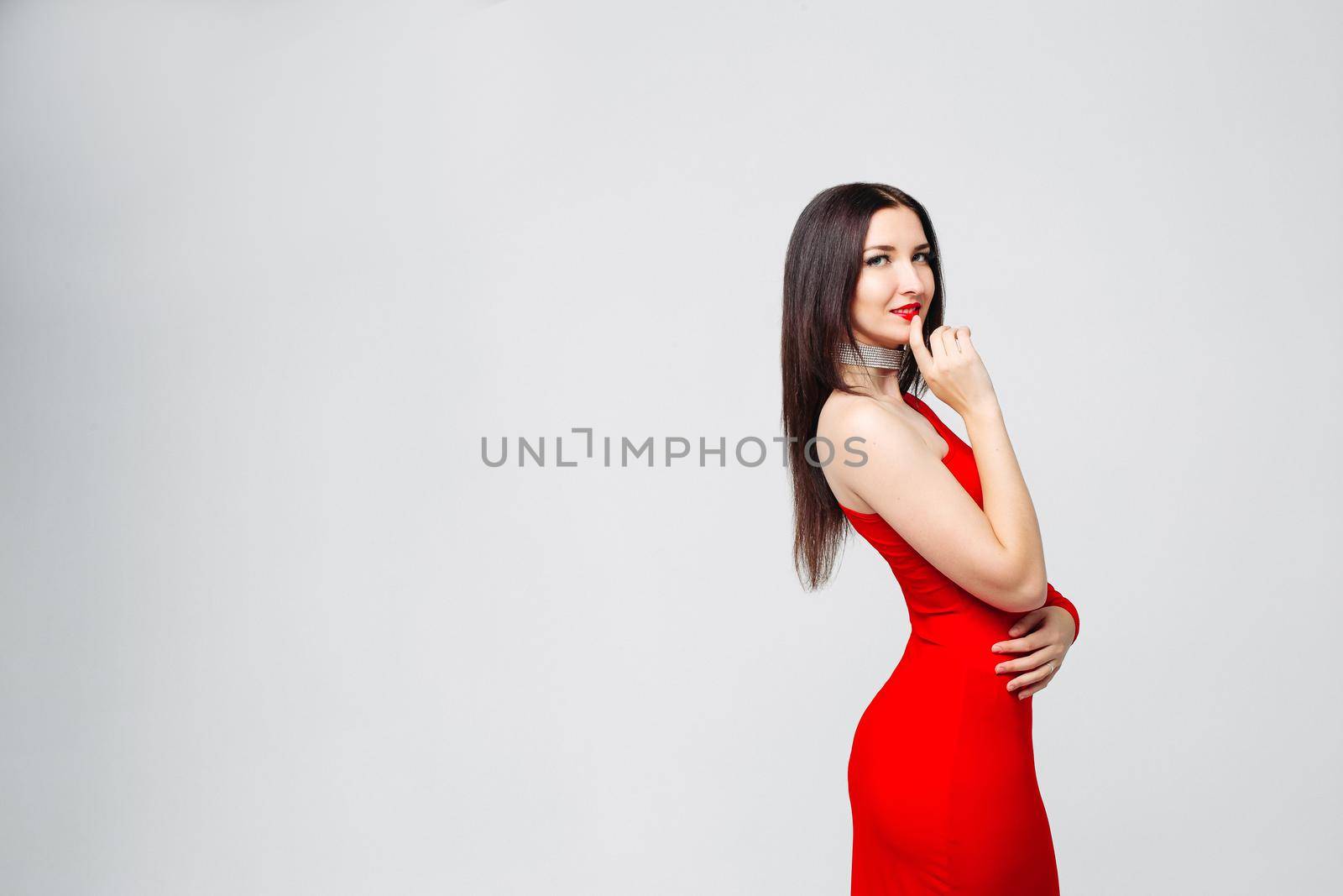 Studio portrait of beautiful sexy woman with long brunette hair wearing bright red dress and make up, embracing herself holding hand at lips over white background. Copyspace.